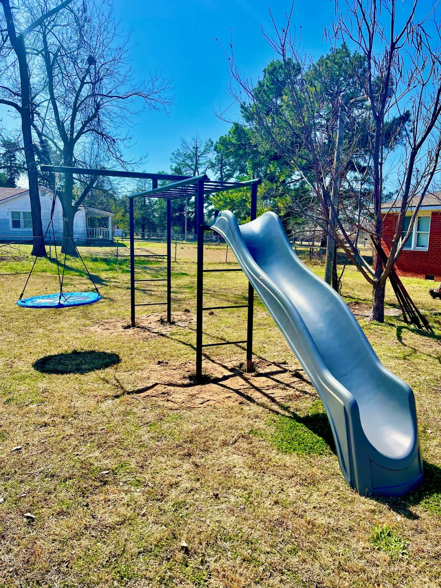 A backyard with a metal playground slide, a round blue swingset, and trees, with a house visible in the background under a clear blue sky.