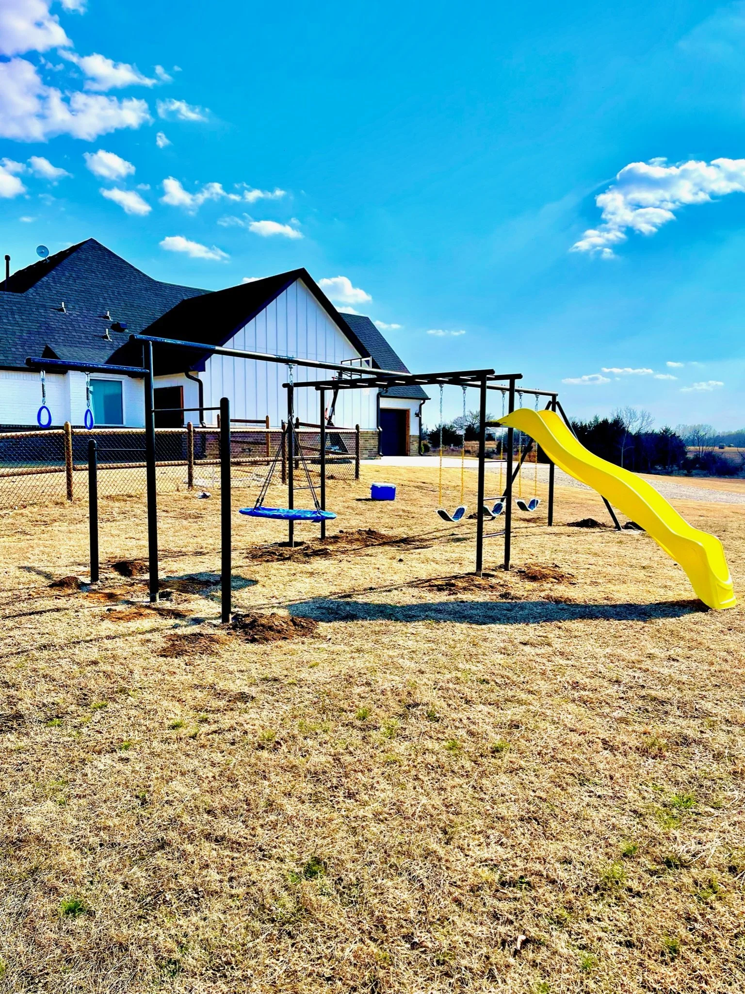Empty playground with swings, slide, and a house in the background under a blue sky with clouds