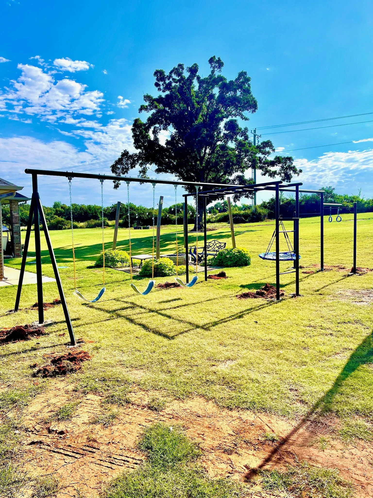 Backyard playground with swings, a seesaw, and a tire swing under a large tree on a sunny day with blue sky and scattered clouds.