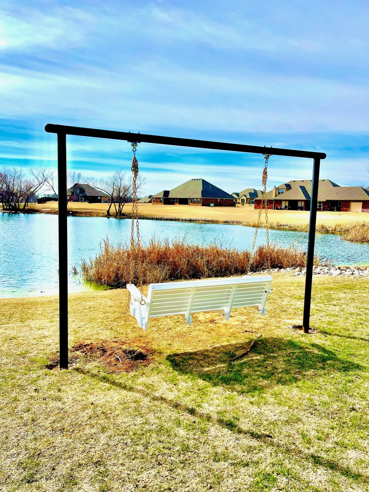 A white porch swing hanging from a metal frame by chains, situated on grass near a small pond with houses in the background under a partly cloudy sky.