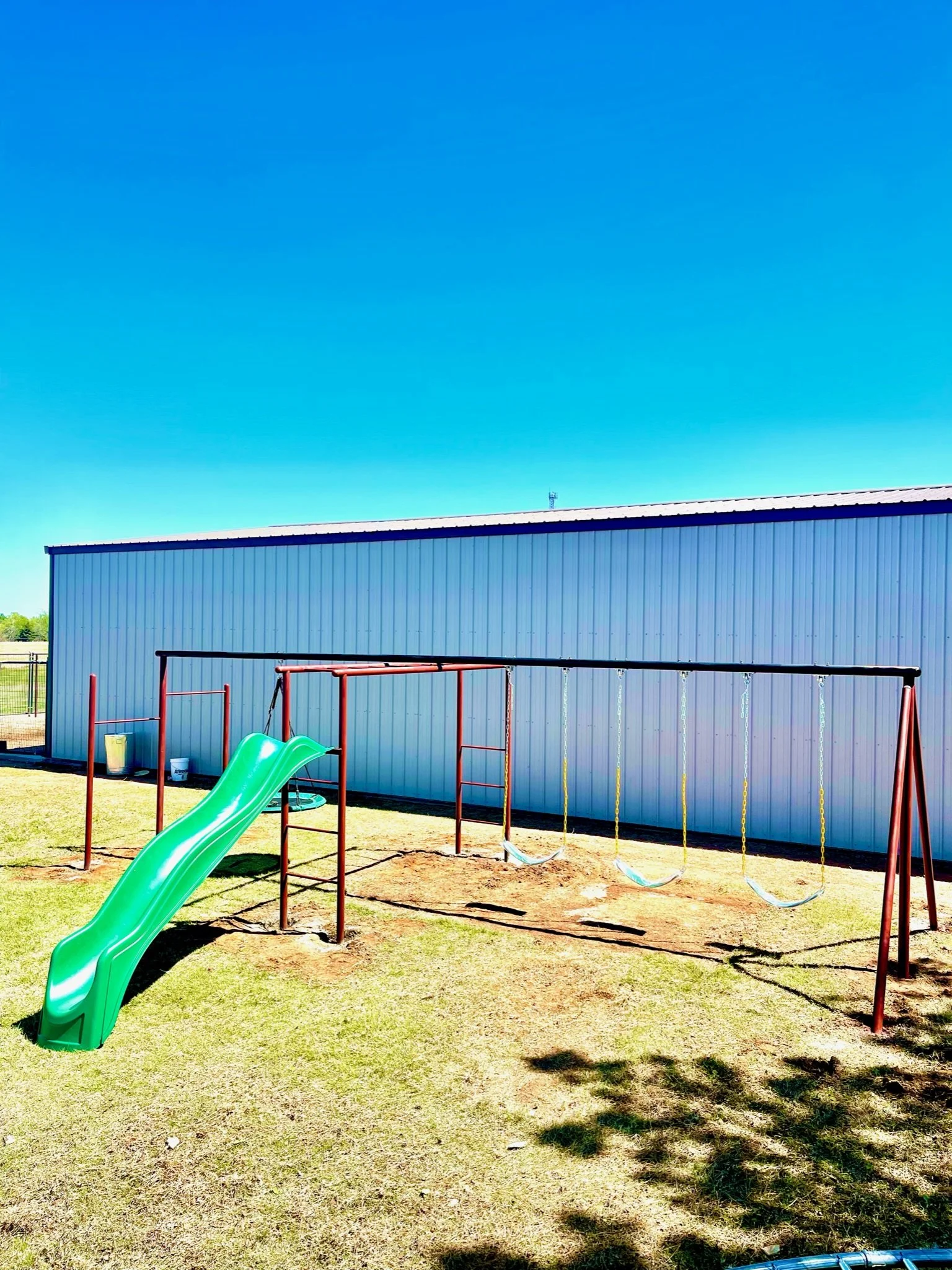 A playground with a green slide and swings in front of a light blue metal building under a bright blue sky.
