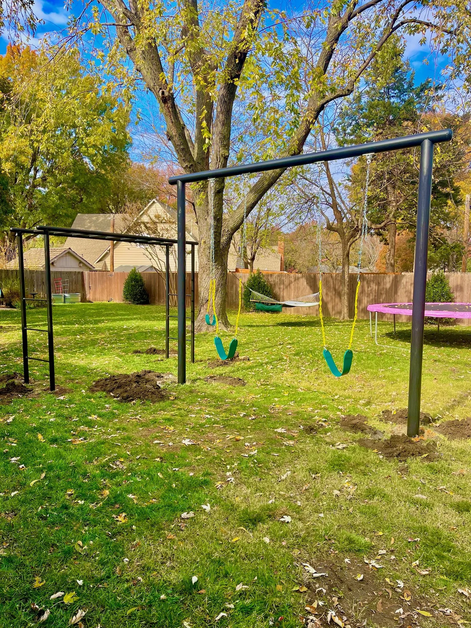 A backyard with a swing set, a hammock, and a trampoline, surrounded by trees with autumn leaves.