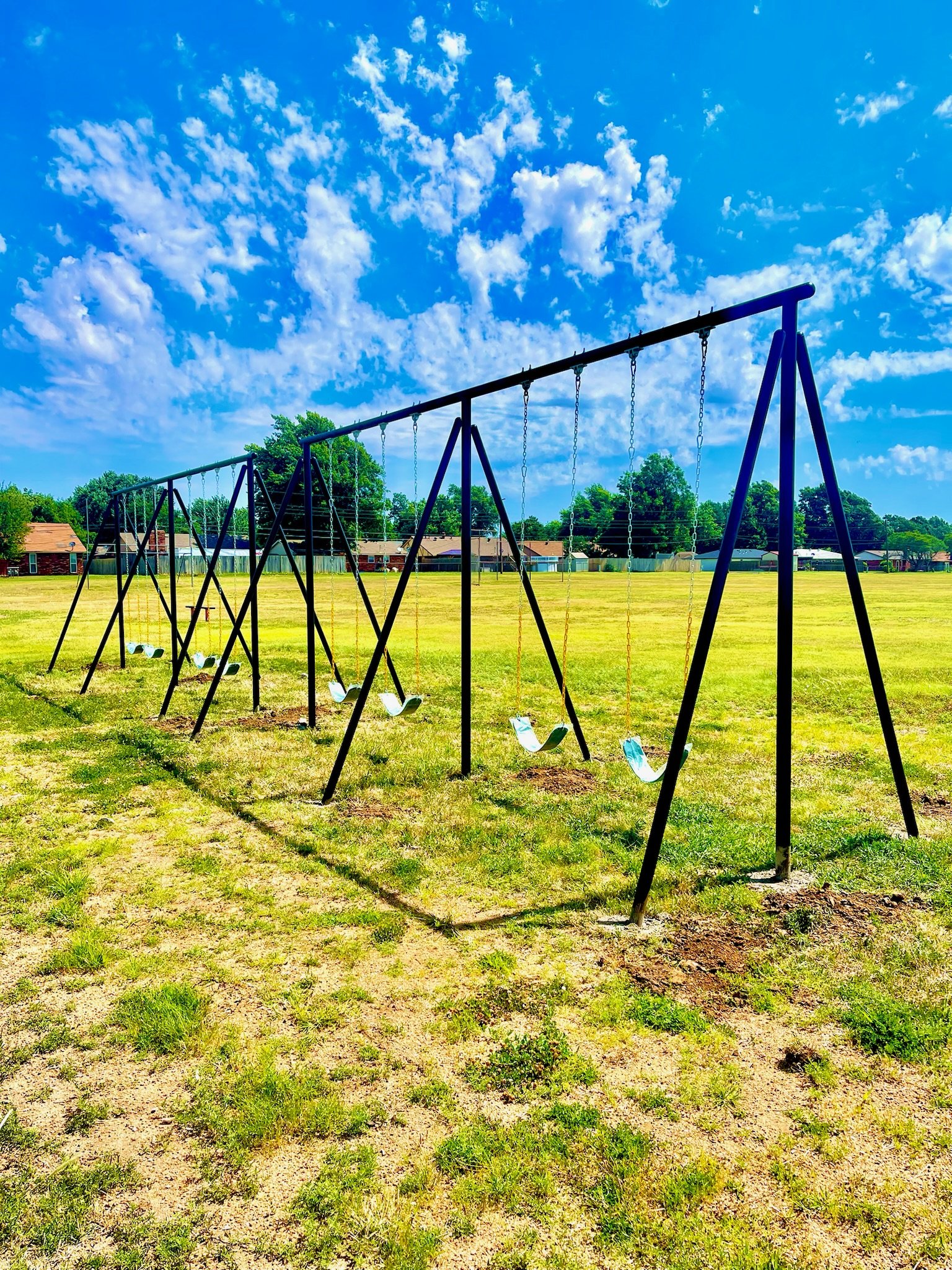 Empty swings set in a grassy field on a bright, sunny day with blue sky and scattered clouds.