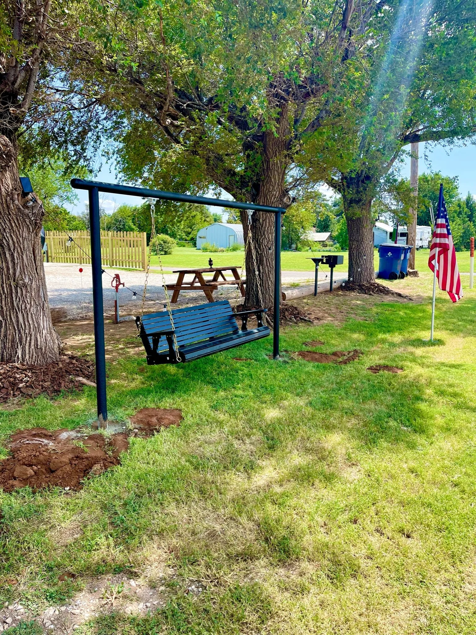 A backyard with a black metal swing seat hanging from a frame, two large trees, a wooden picnic table, an American flag on a pole, a blue trash bin, a barbecue grill, and a white shed in the background on a sunny day.