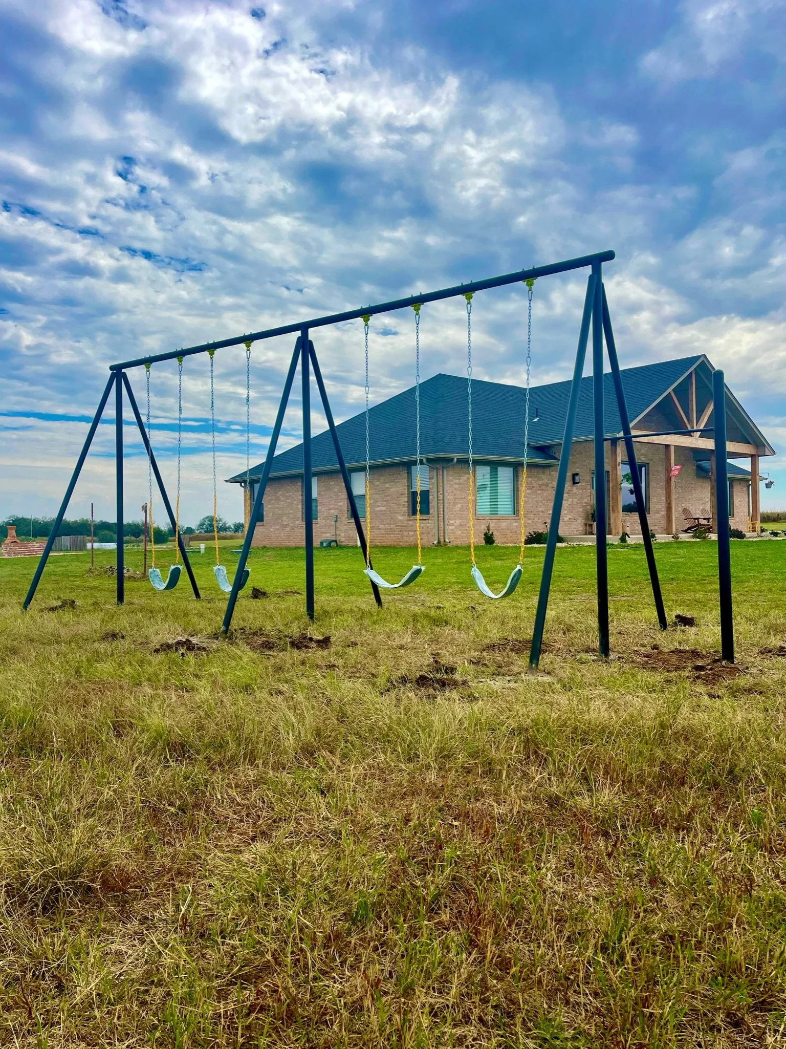 A playground swing set with five swings in front of a brick house under a partly cloudy sky.