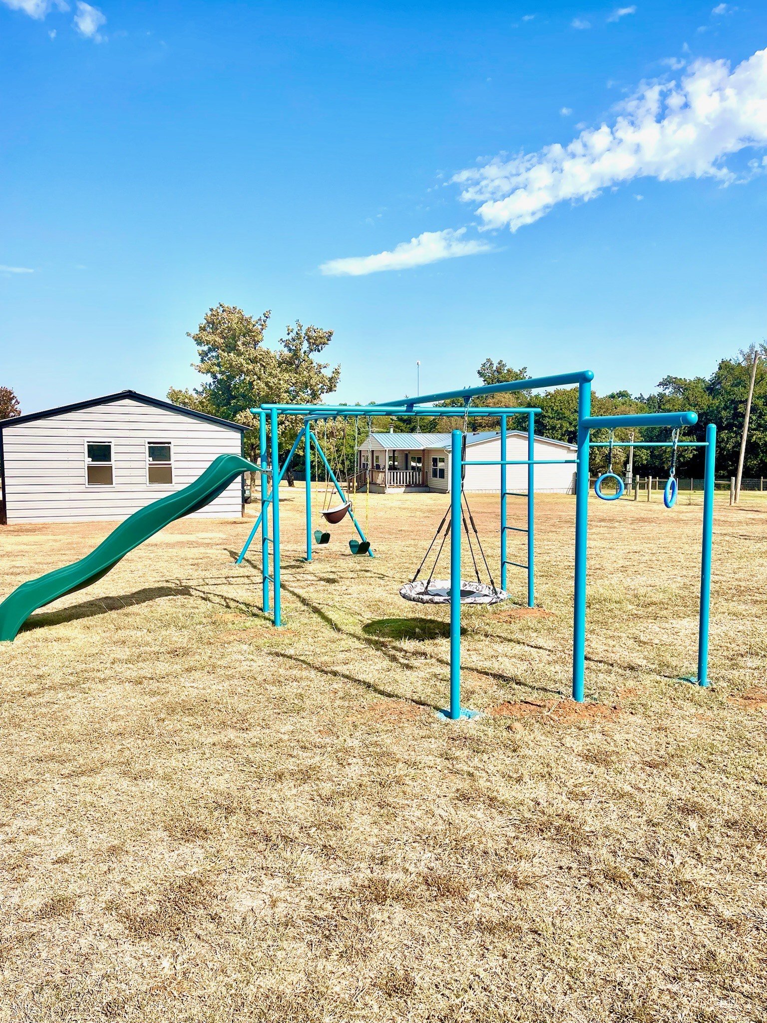 Blue playground set with slide, swings, and gym rings in a grassy yard with a shed and house in the background under a partly cloudy sky.