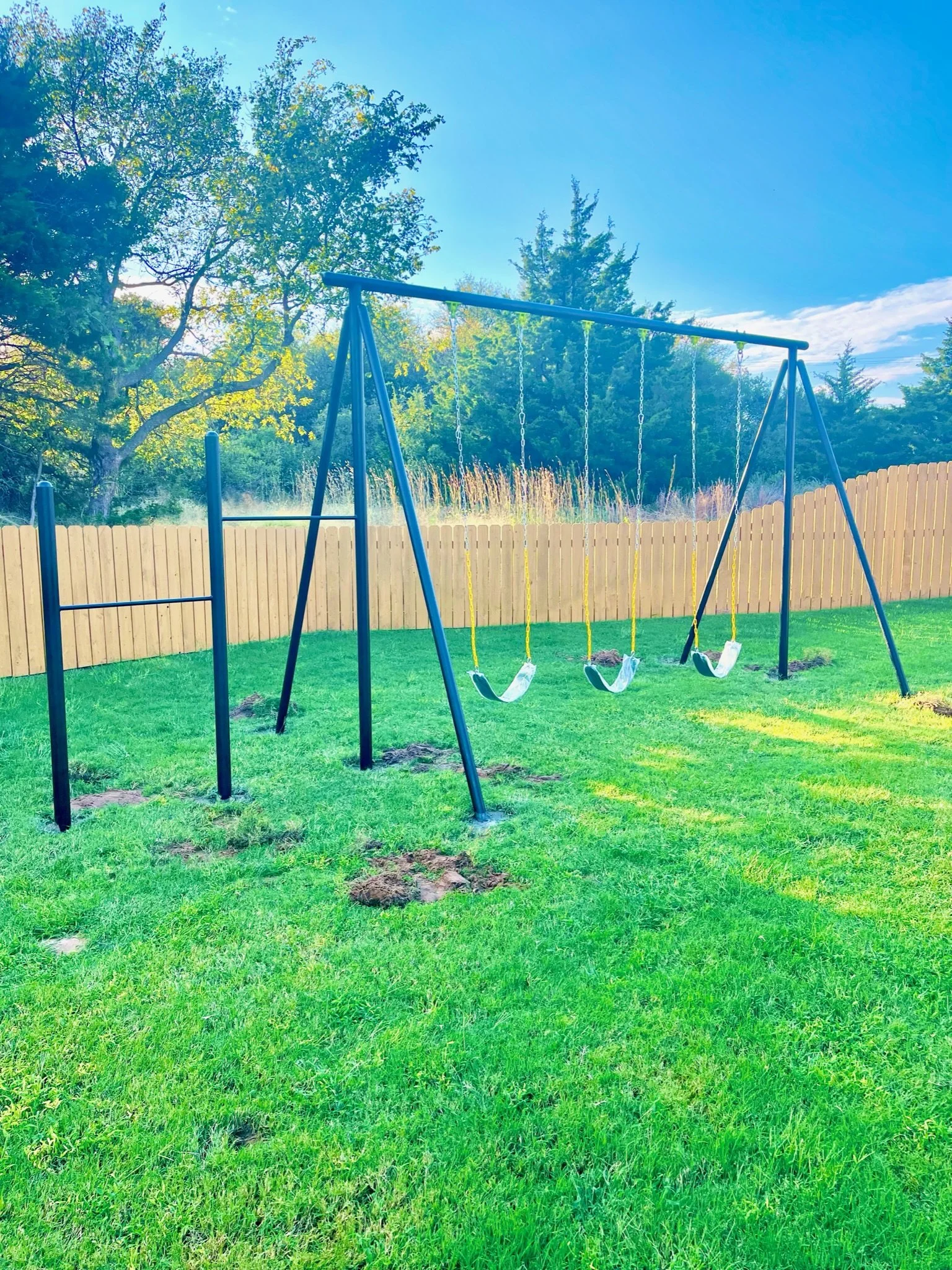 A playground swing set with four swings in a grassy backyard, surrounded by a wooden privacy fence and trees in the background under a partly cloudy sky.