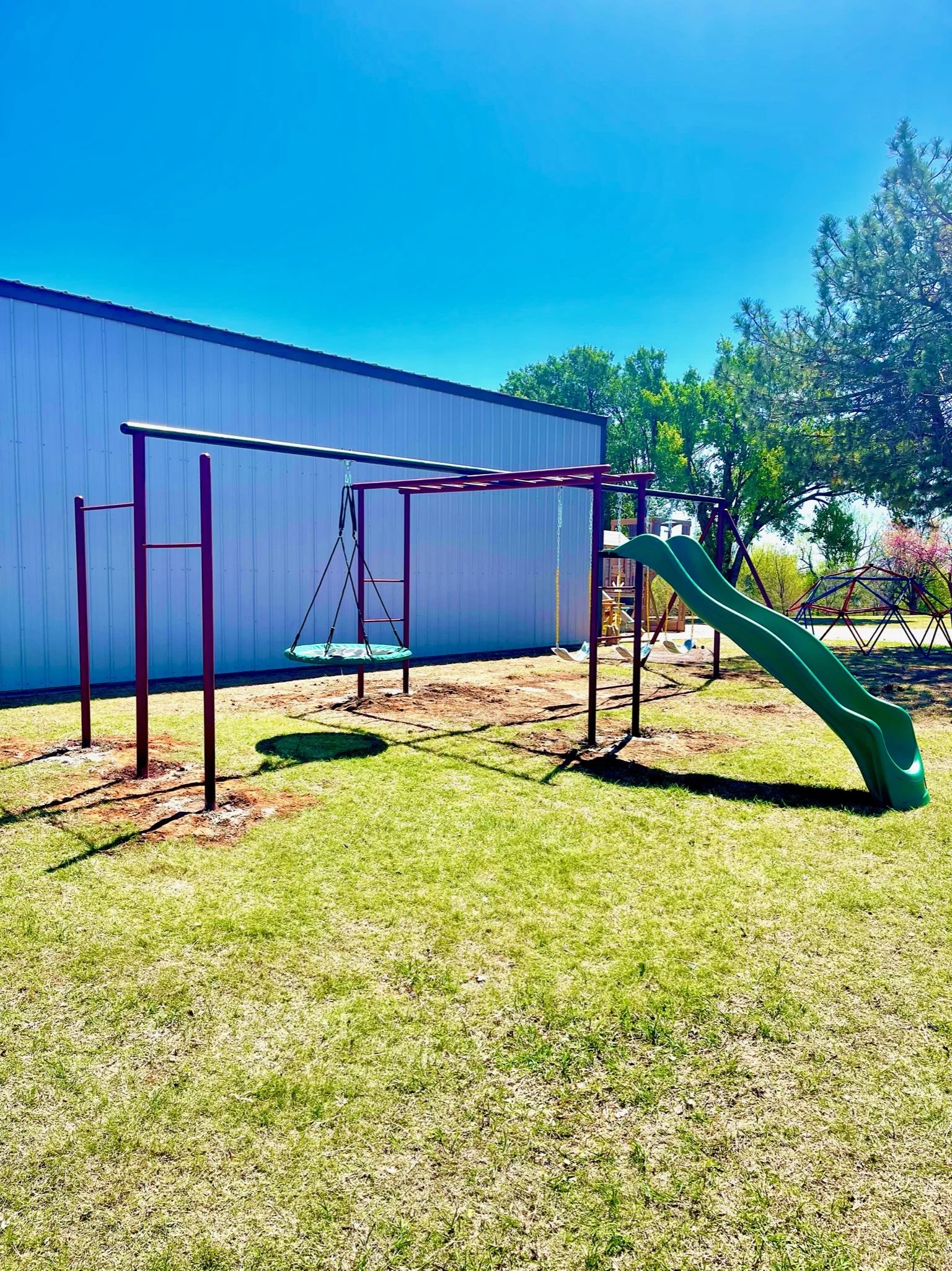 Children's playground with swing set, slide, and climbing structure in a grassy yard under a clear blue sky.