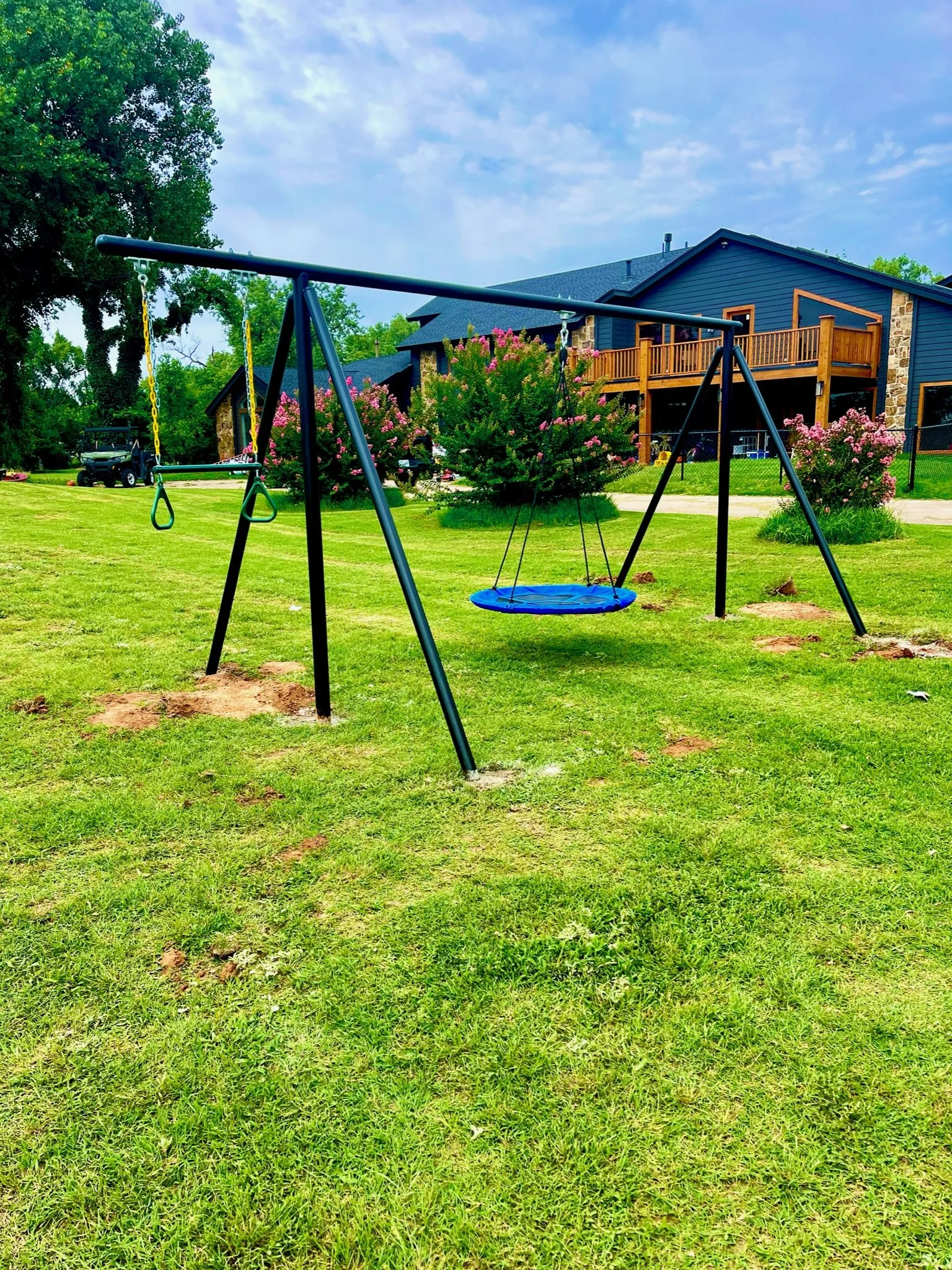 Playground swing set on green grass with a house and pink flowering bushes and trees in the background.