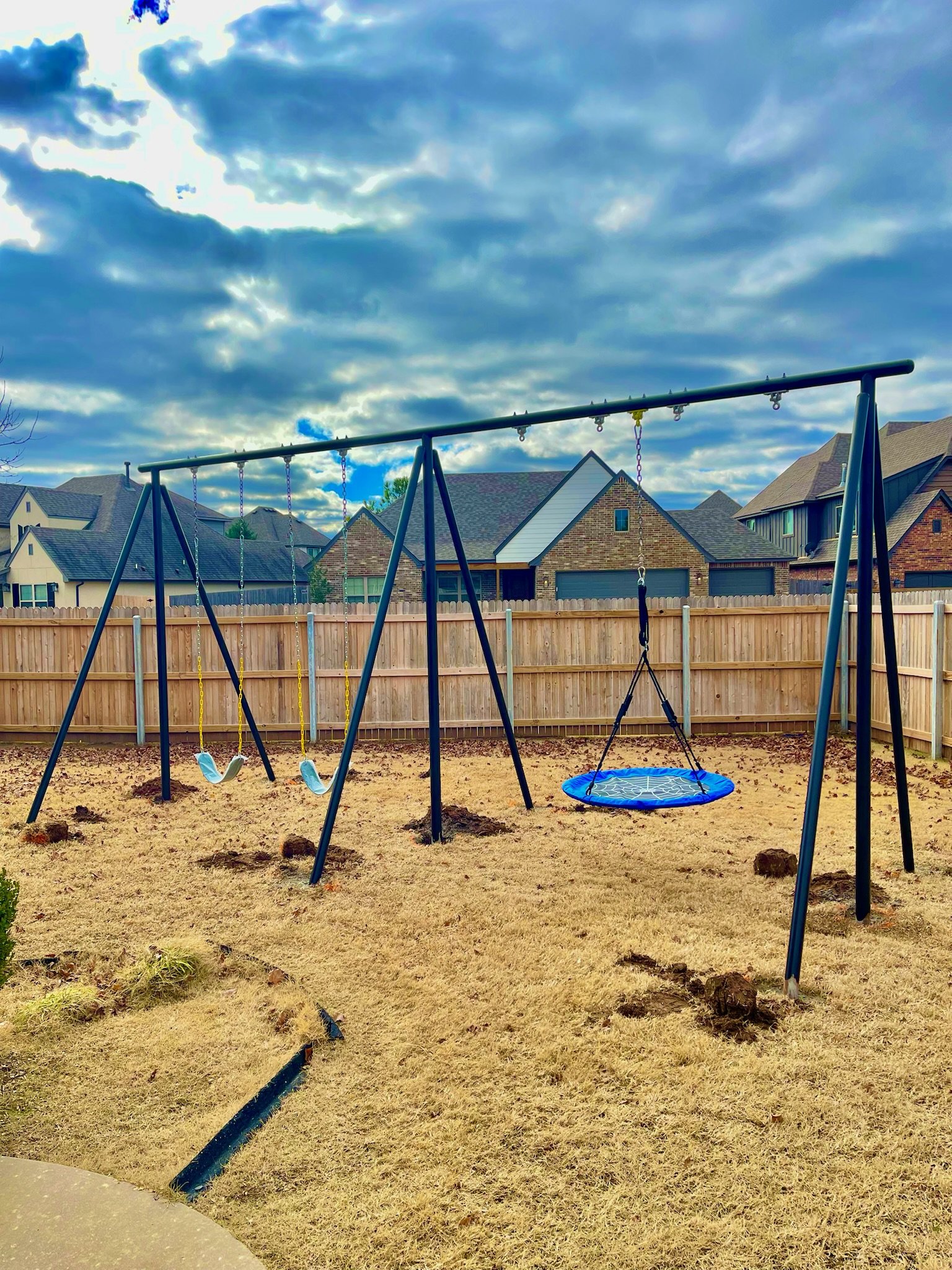 A backyard playground set featuring two swings and a basket swing, set on a grassy yard with a wooden fence and suburban houses in the background under a cloudy sky.