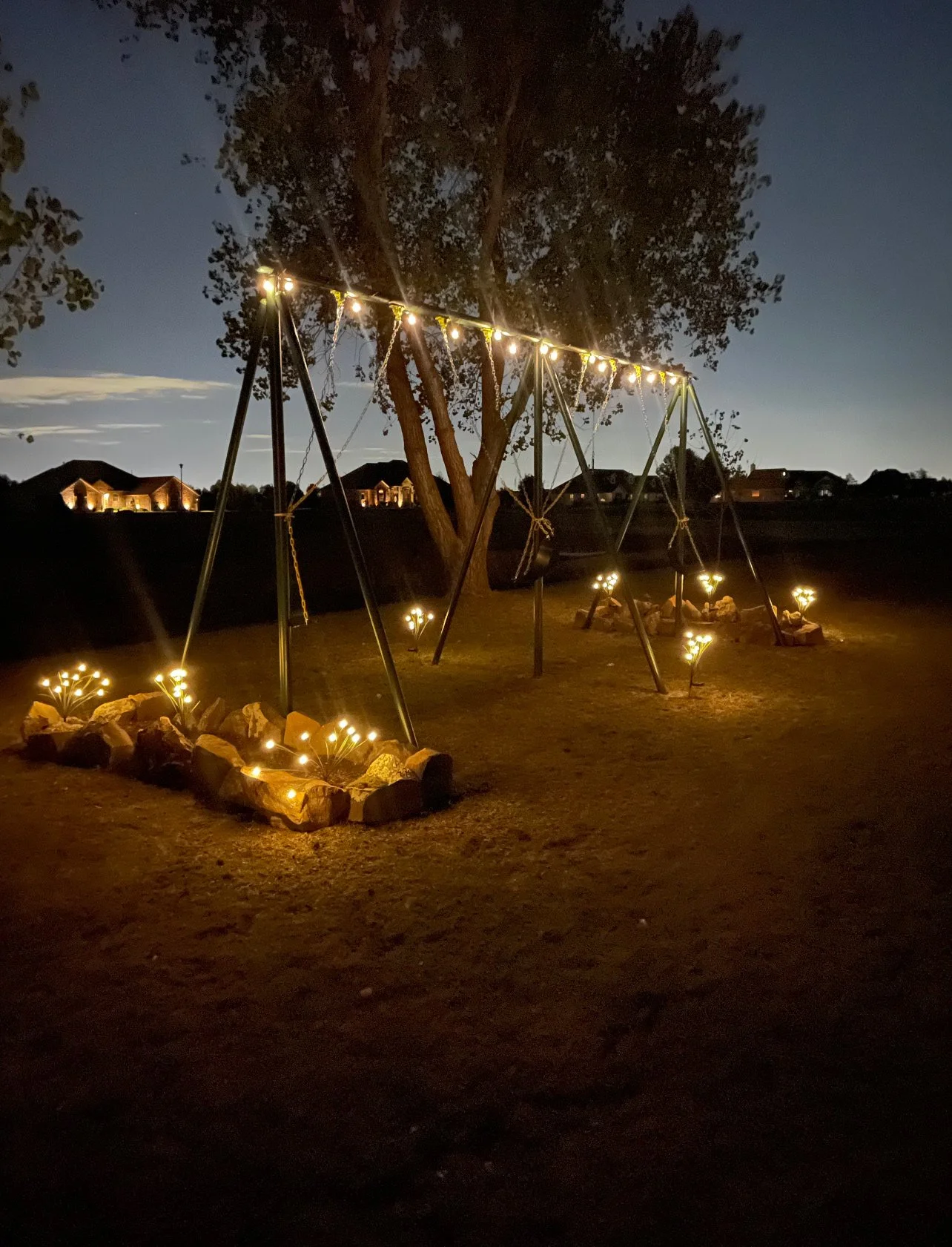 A tree with a swing set hanging from it, decorated with string lights. Small flower-shaped lights are placed around the grass, creating a cozy nighttime outdoor setting.