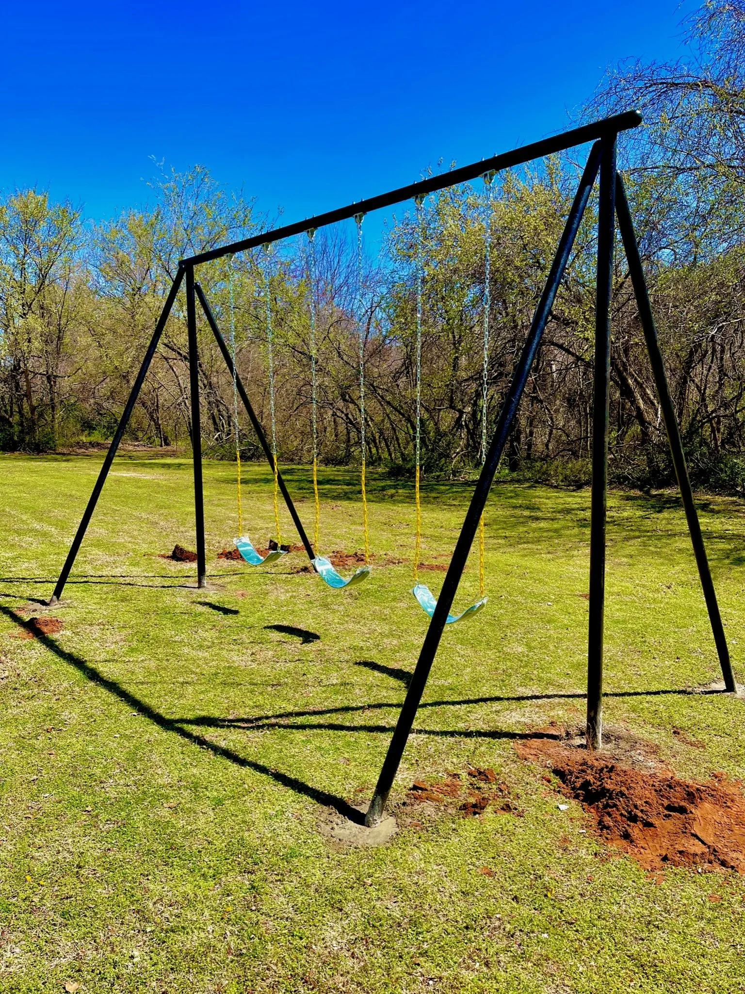 A black metal swing set with four yellow swings on a grassy field, with trees and a bright blue sky in the background.