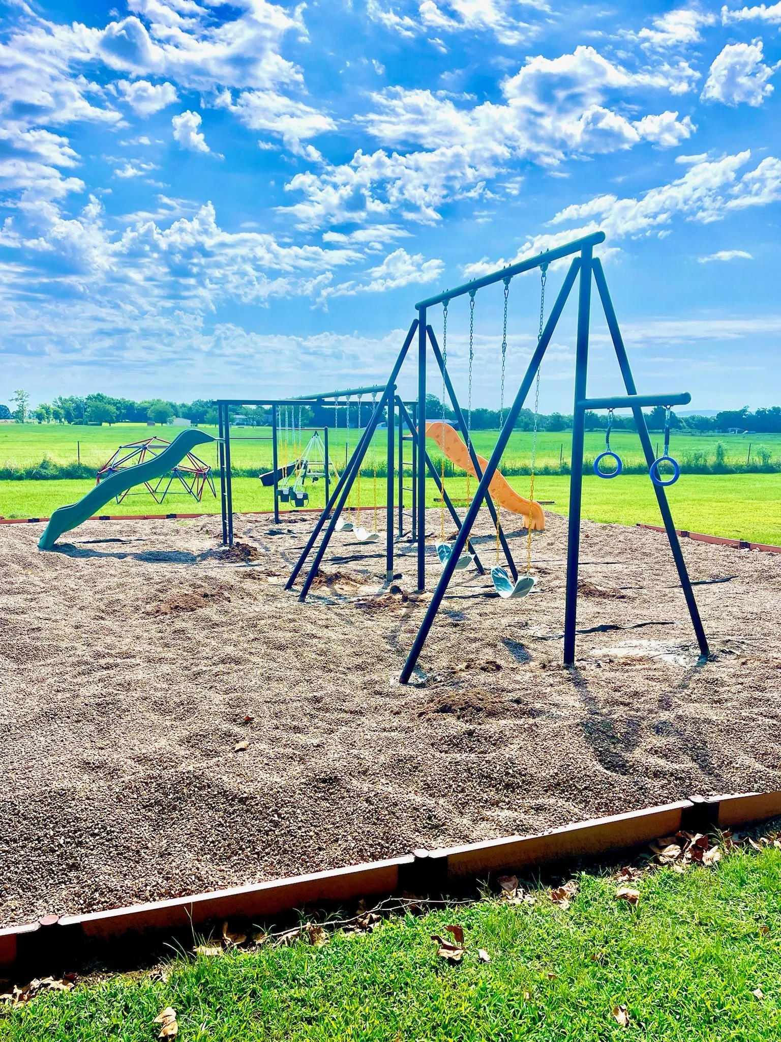 Childrens playground with swings, slides, and climbing structures on a sandy area with green fields and a blue sky with clouds in the background.