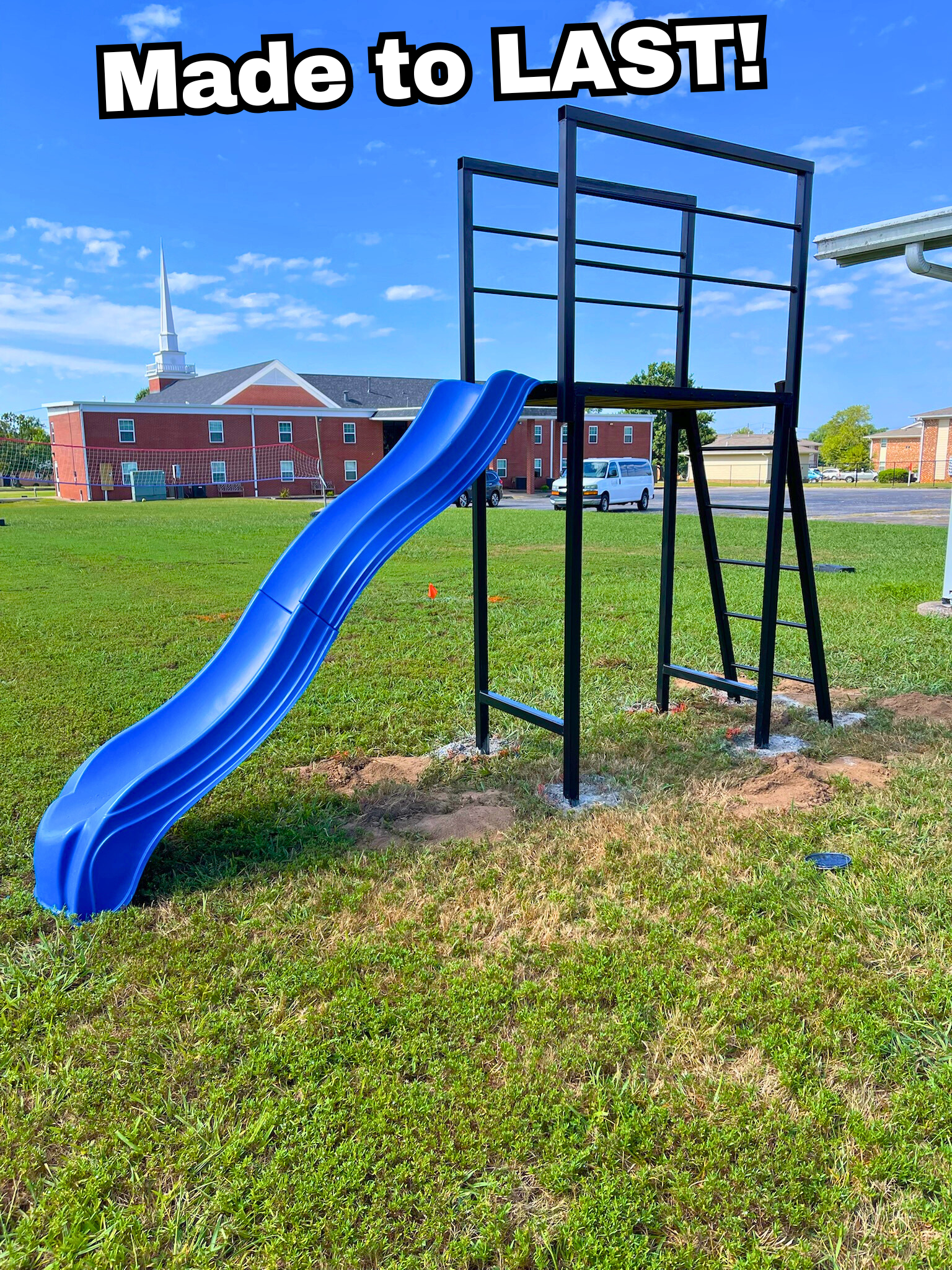 A playground slide with a blue slide and black metal frame on a grassy field, with a brick building and church steeple in the background, and the text 'Made to LAST!' at the top of the image.