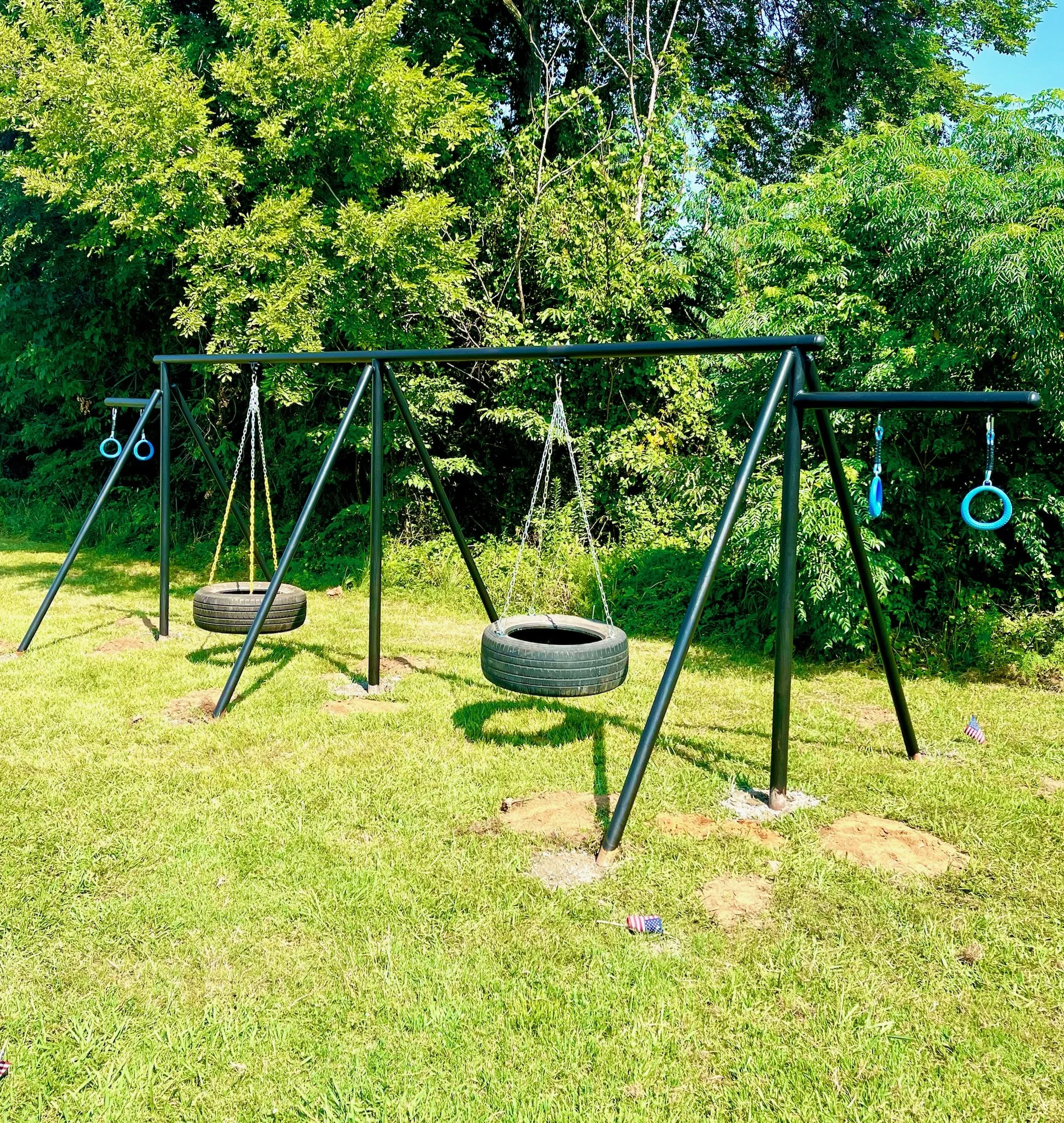 Outdoor playground set with two tire swings and two blue gymnastic rings on a grassy field with trees in the background