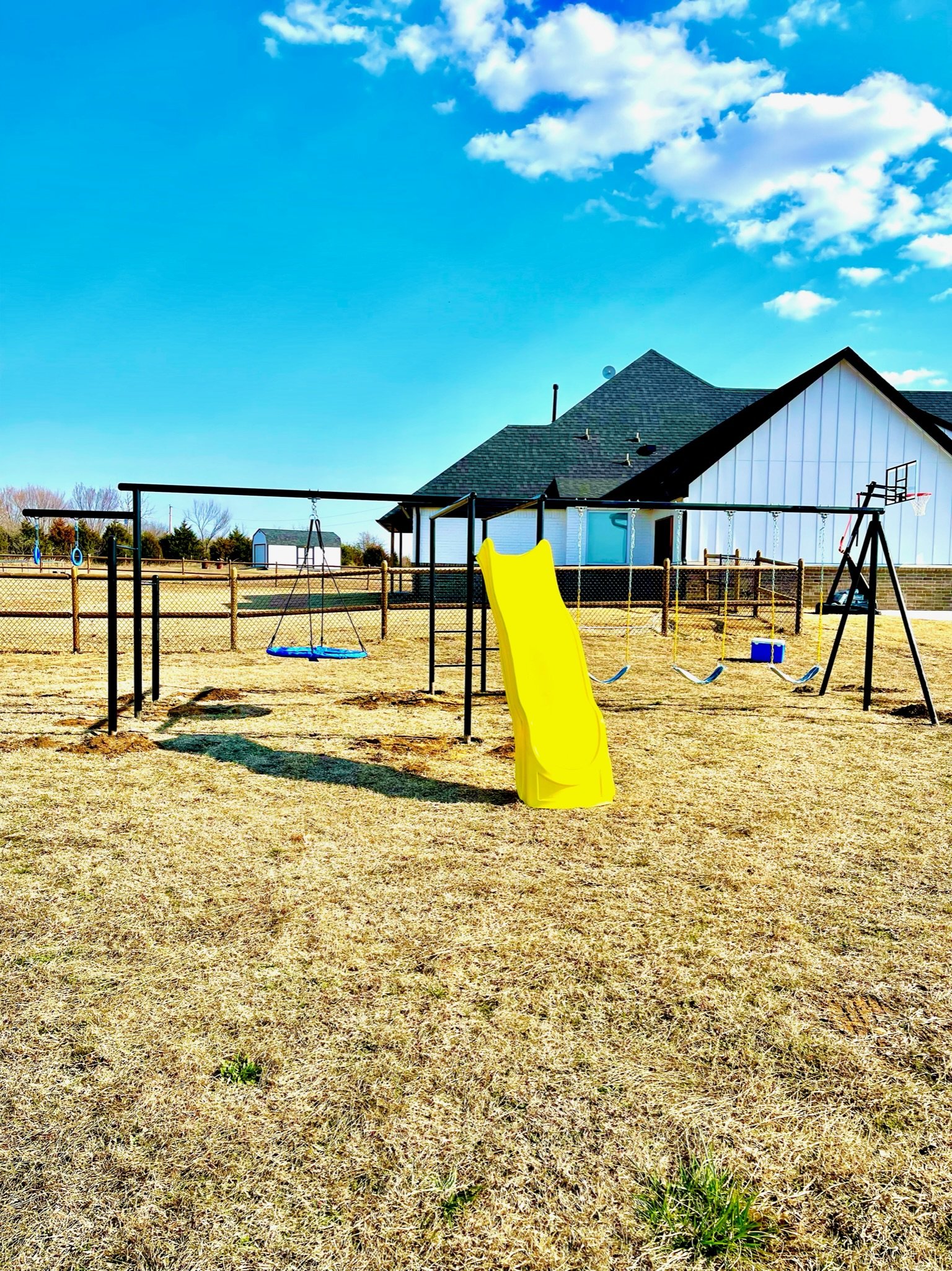A playground with a yellow slide, swings, a basketball hoop, and a fenced yard in front of a house with a gabled roof, under a blue sky with a few clouds.