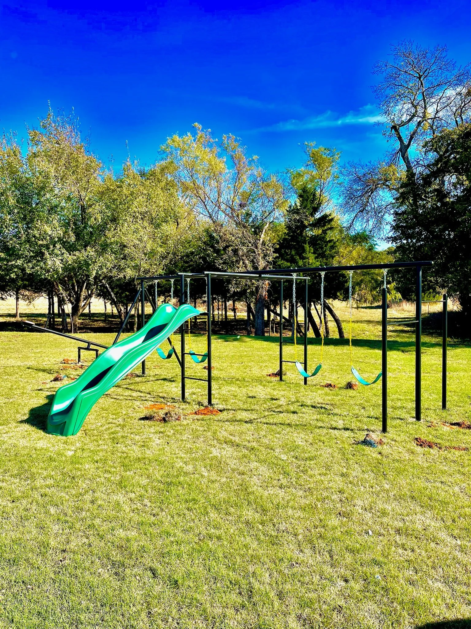 A playground with a green slide and several swings set in a grassy field under a bright blue sky, surrounded by trees.