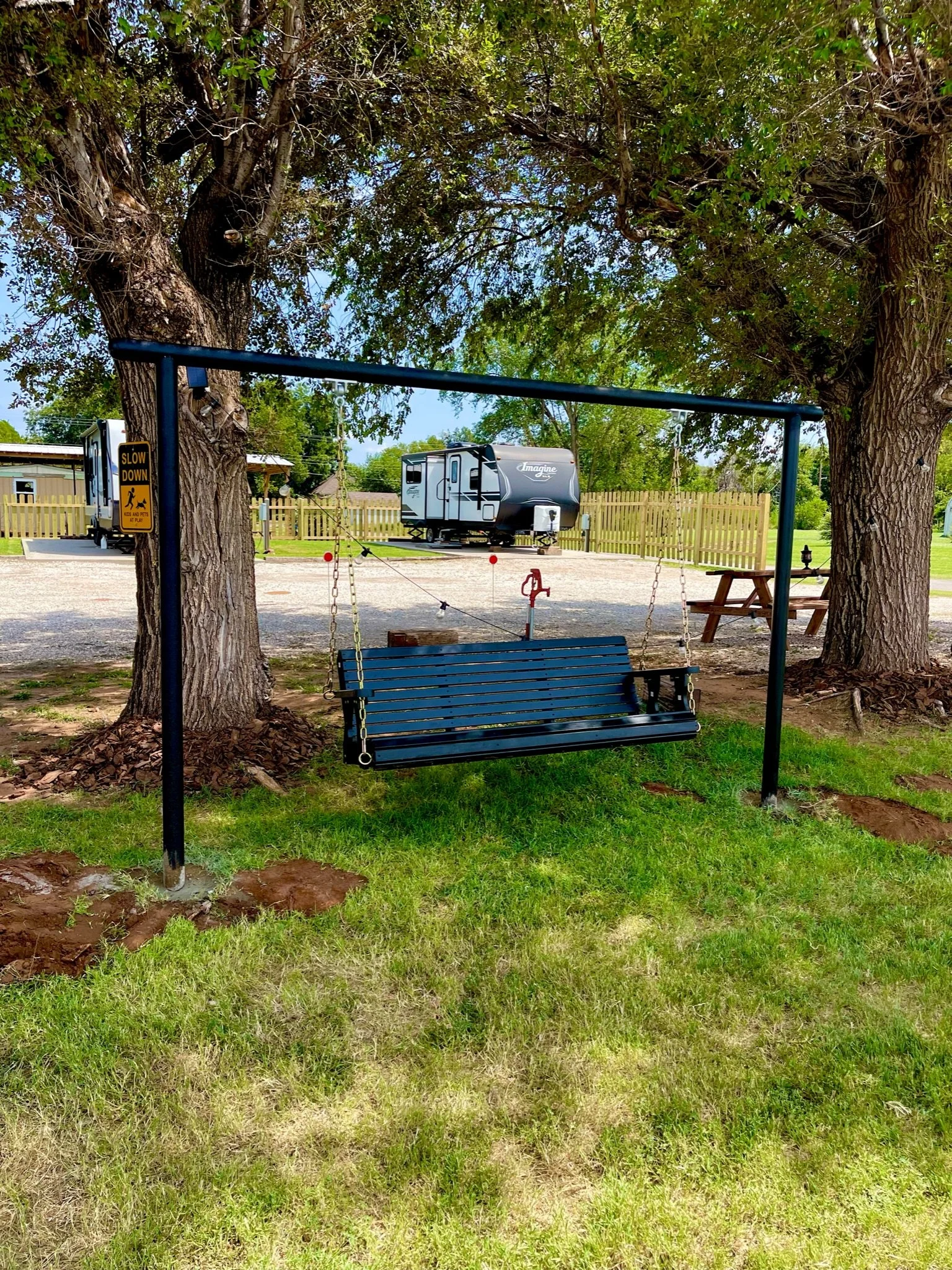 A black metal swing hanging from a blue frame, set under a large tree with a park or campsite area in the background, including a camper trailer and a picnic table.