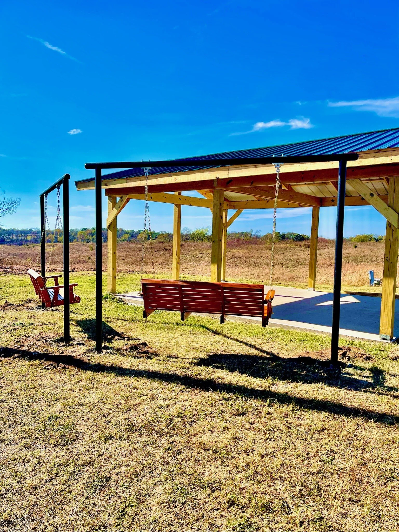 A swing set with two red swings, a wooden porch with a covered roof, placed on a grassy area with a field in the background under a bright blue sky.