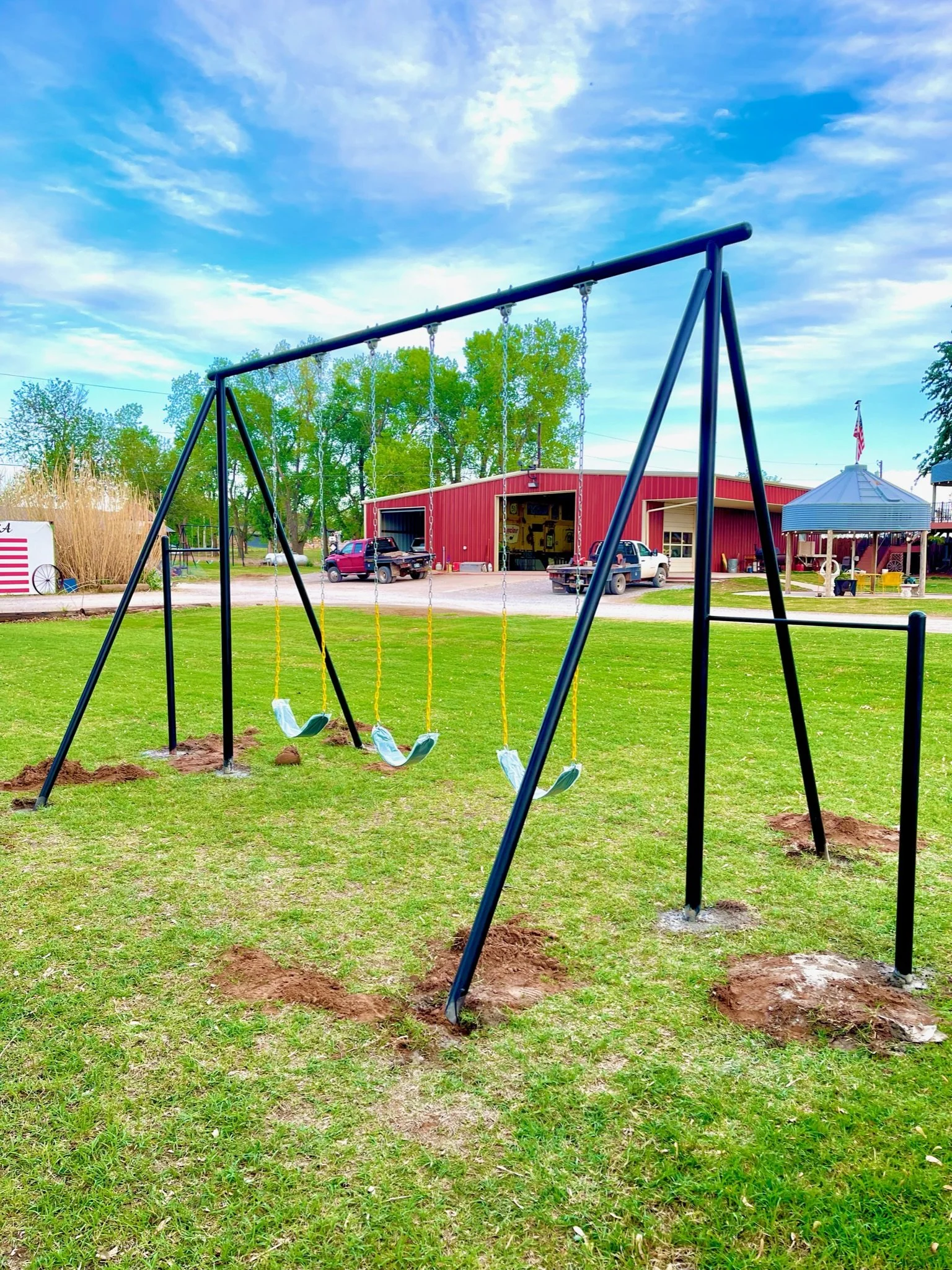 Empty swing set on grassy field with a red barn, vehicles, and trees in the background under a partly cloudy sky.