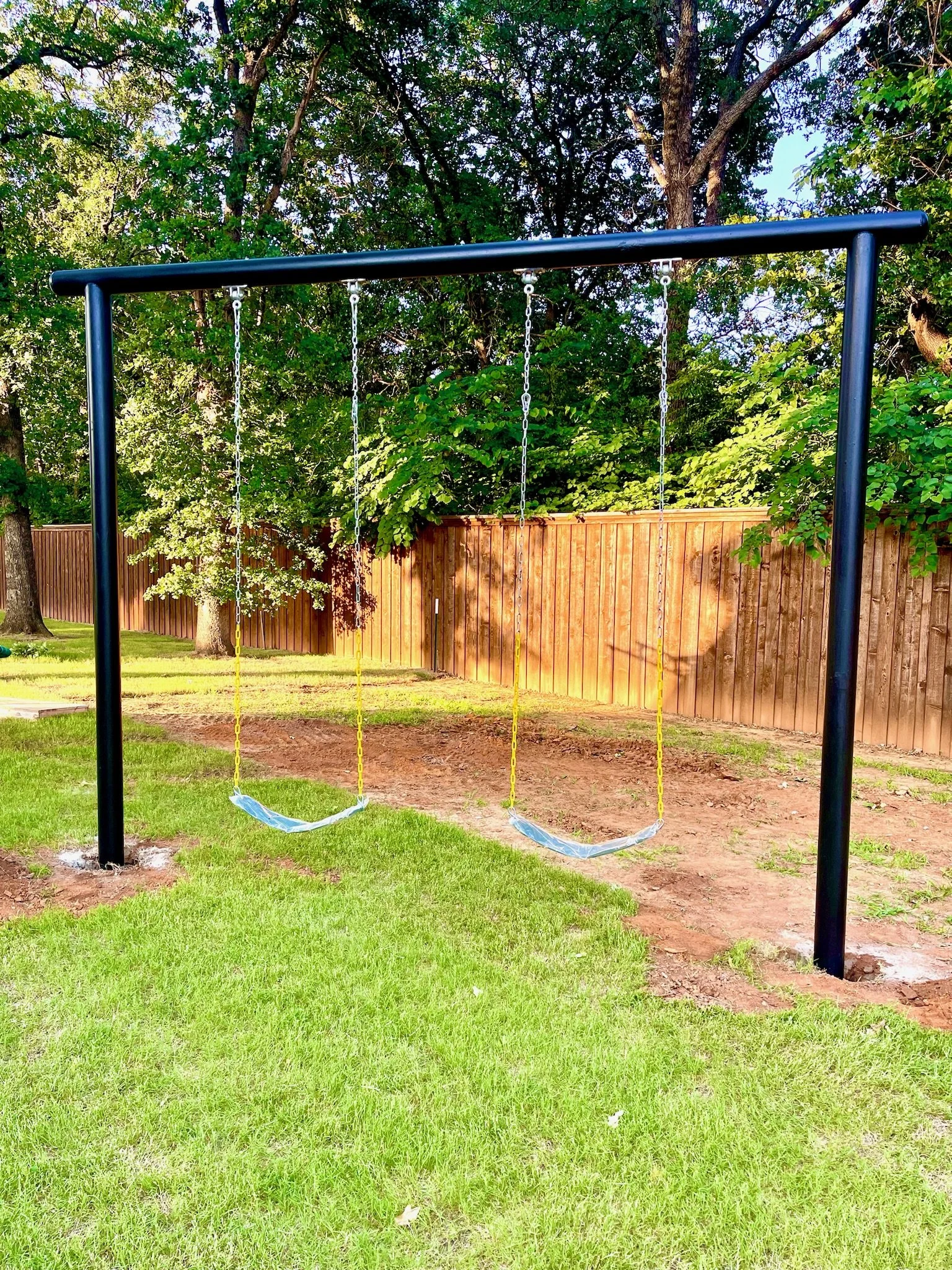 Empty swings on a black metal swing set in a backyard with green grass, trees, a wooden fence, and dirt patches.