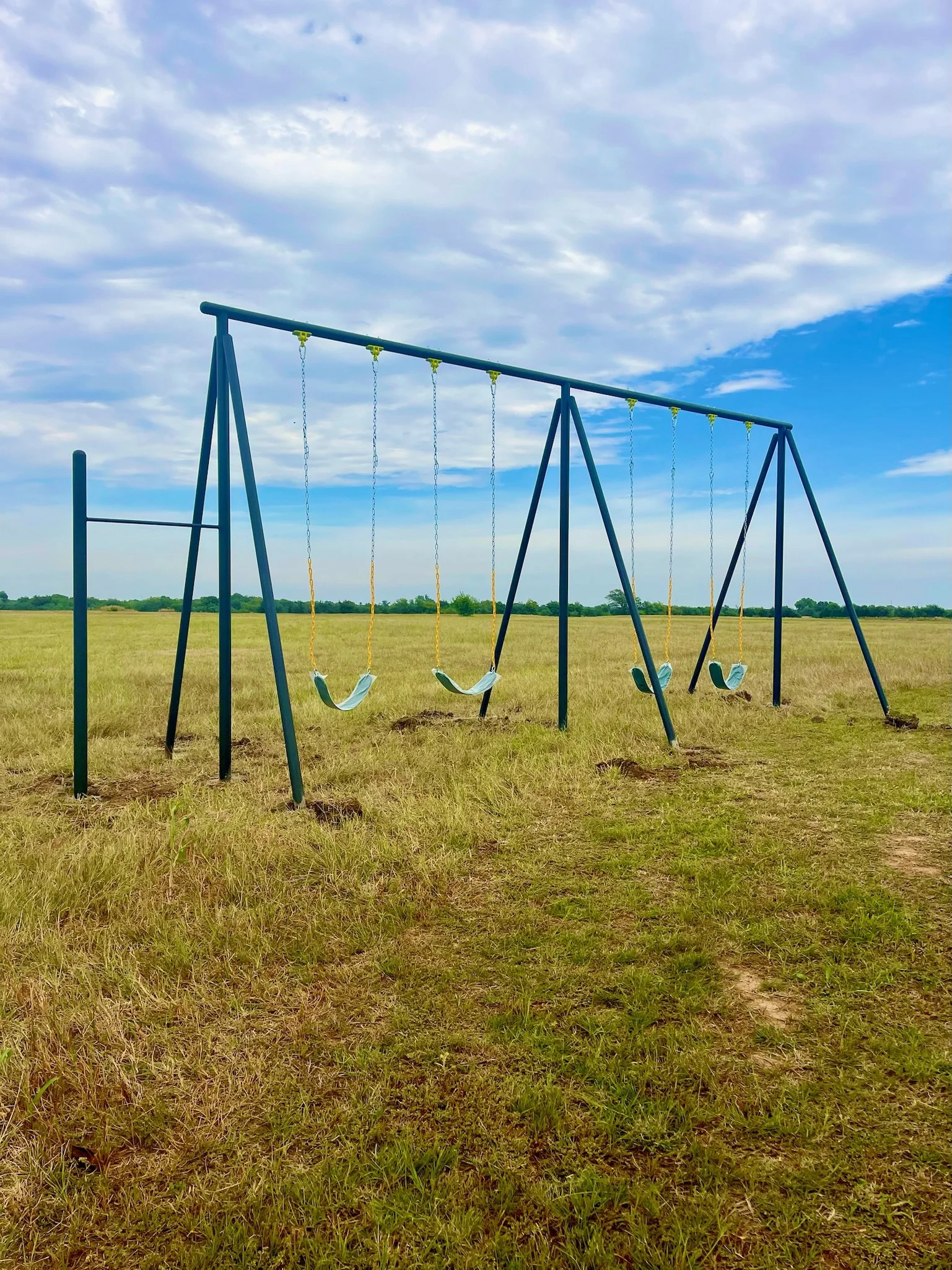 Empty swing set with four swings on a grassy field under cloudy sky.
