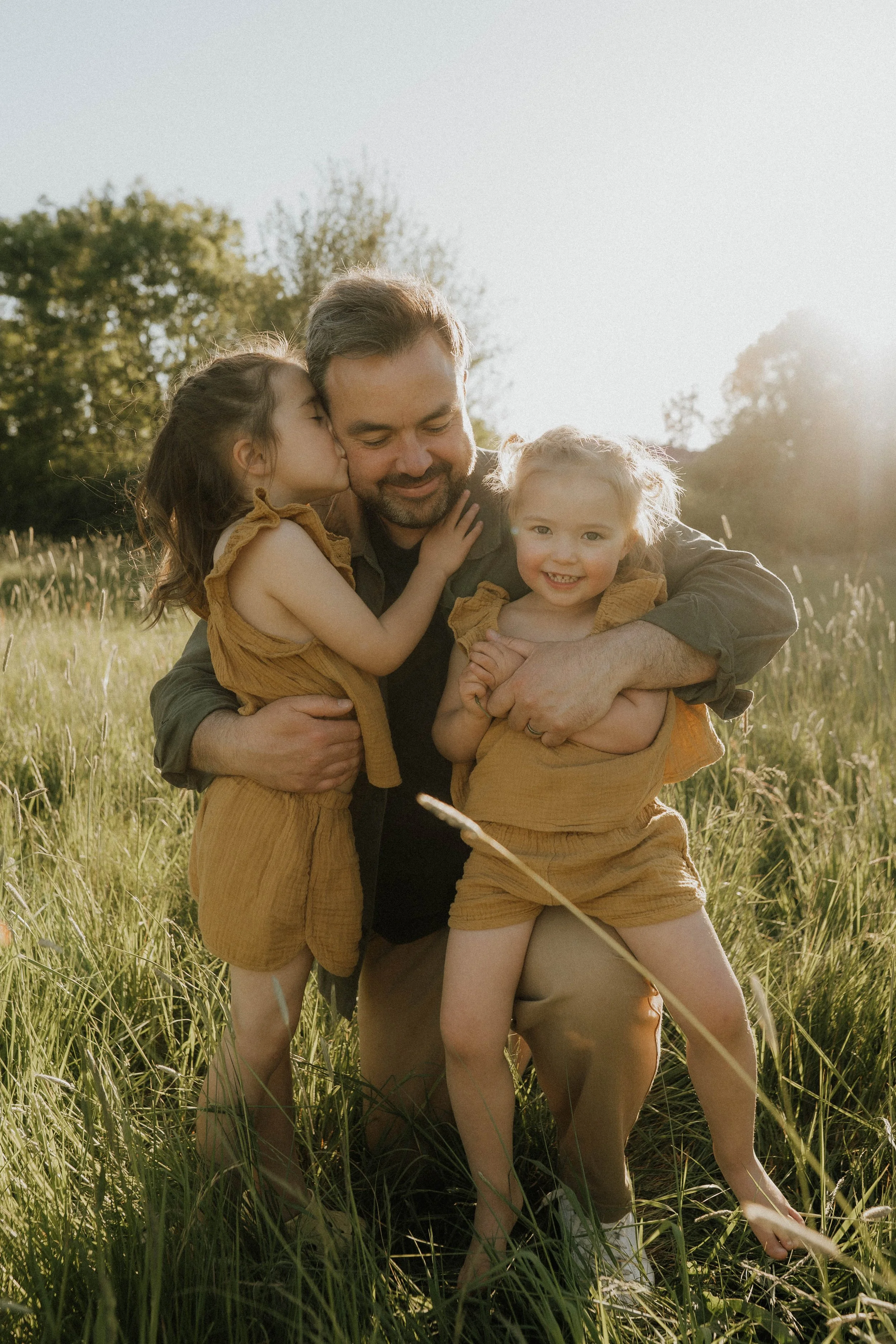 Image of dad and his two young girls in an evening sunlit meadow on their family shoot with Didsbury family photographer Copper + Olive Photo