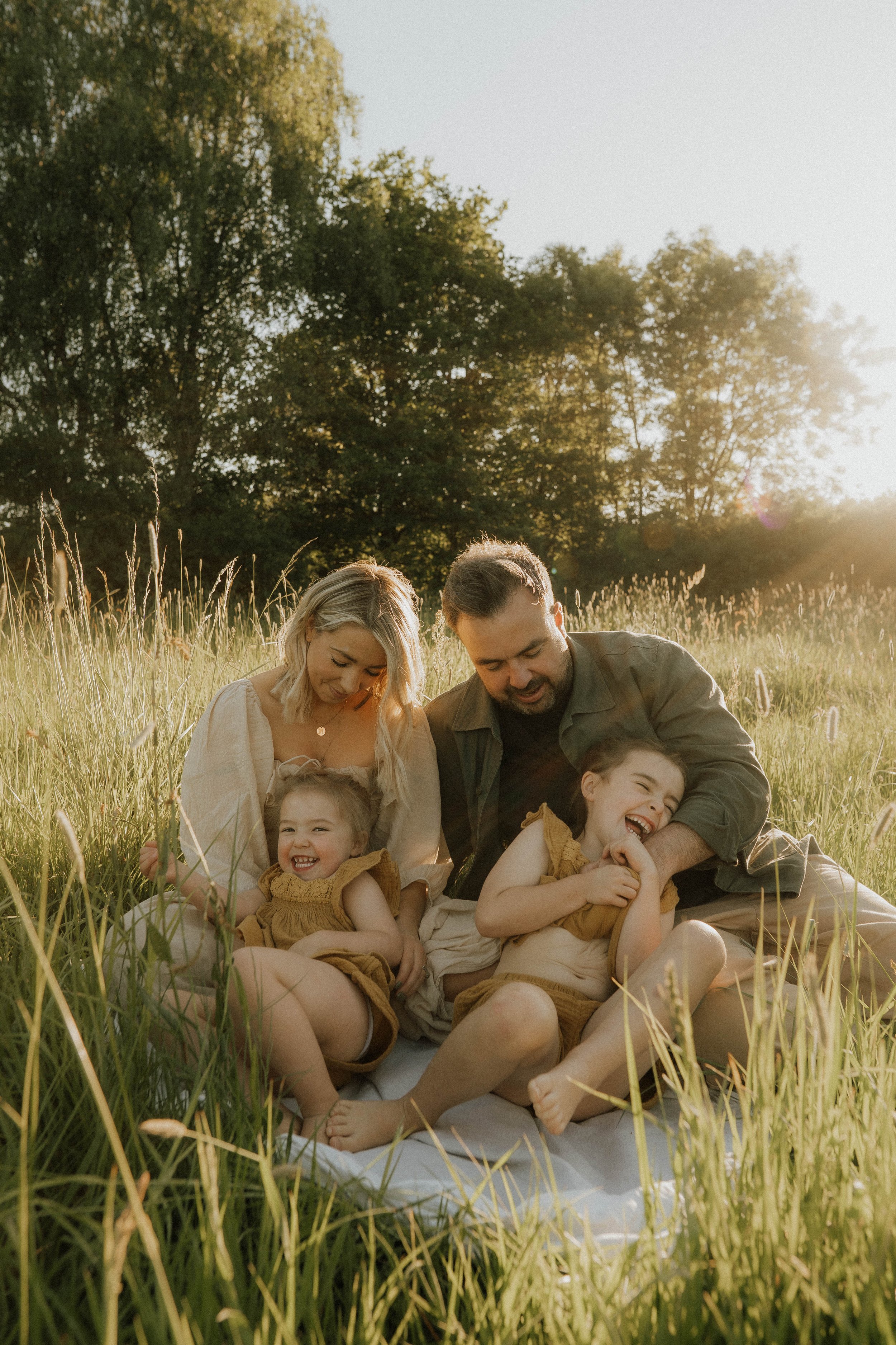 Image of full family of four, sat down together in an evening sunlit meadow, laughing on their family shoot with Didsbury family photographer