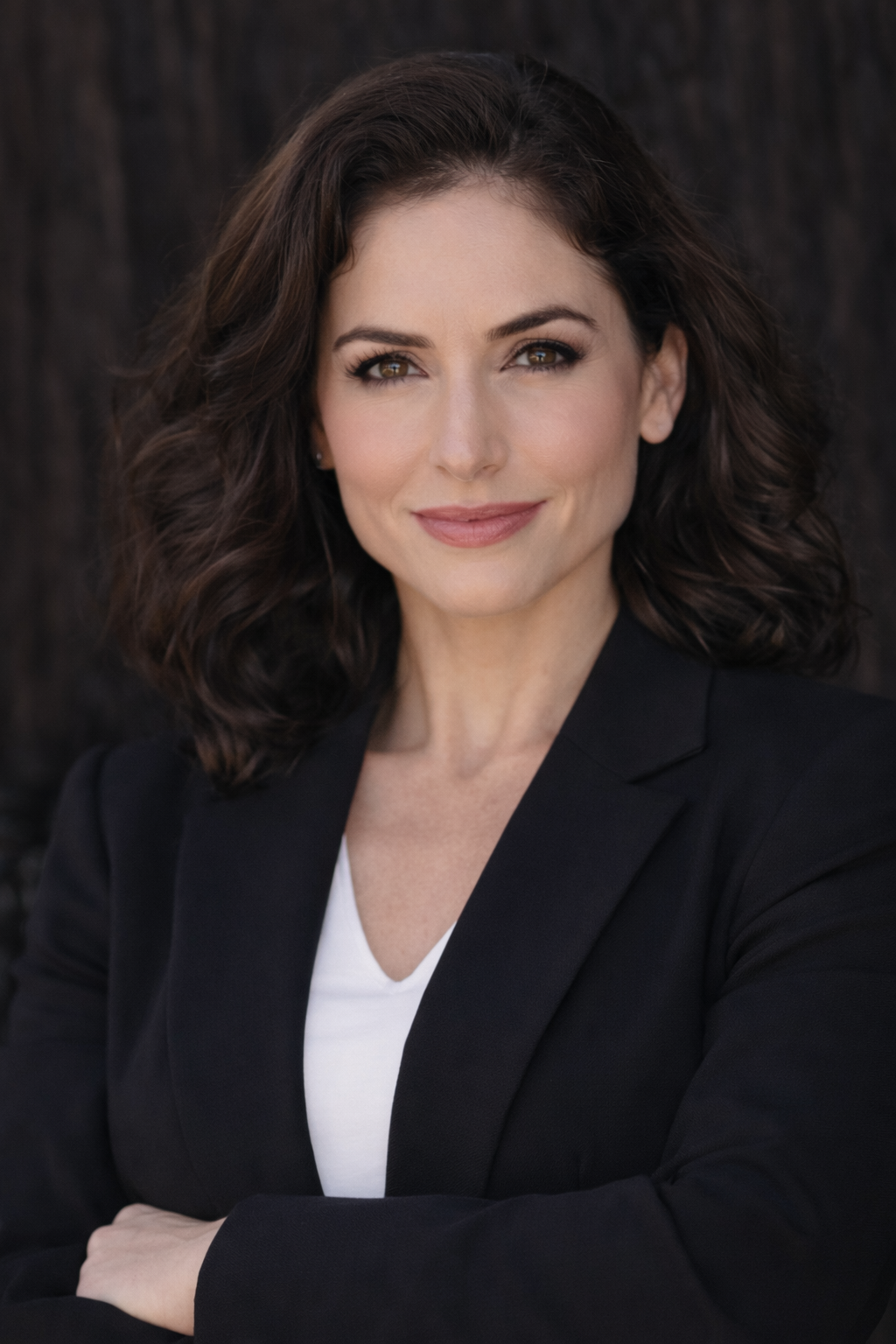 A woman with dark wavy hair and light skin, dressed in a black blazer over a white top, smiling confidently against a dark background.