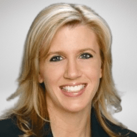 A woman with blonde hair, smiling, wearing a dark blazer, against a neutral background.