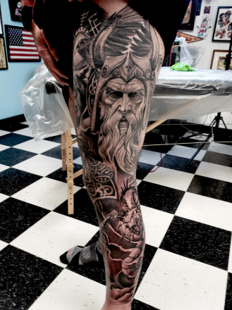 Close-up of a person's leg with an intricate black and gray tattoo of a Viking warrior with a helmet and long beard, set against a tattoo shop background.