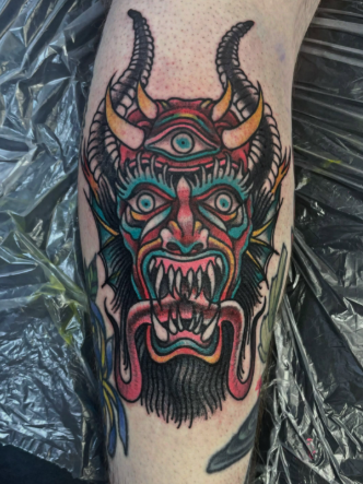 Colorful tattoo of a demon face with horns, multiple eyes, sharp teeth, and flames or tentacles around it, on a person's leg.