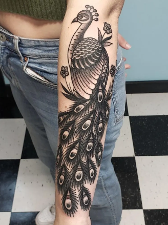 A person's arm with a black and gray tattoo of a peacock with detailed feathers, set against a light blue background.
