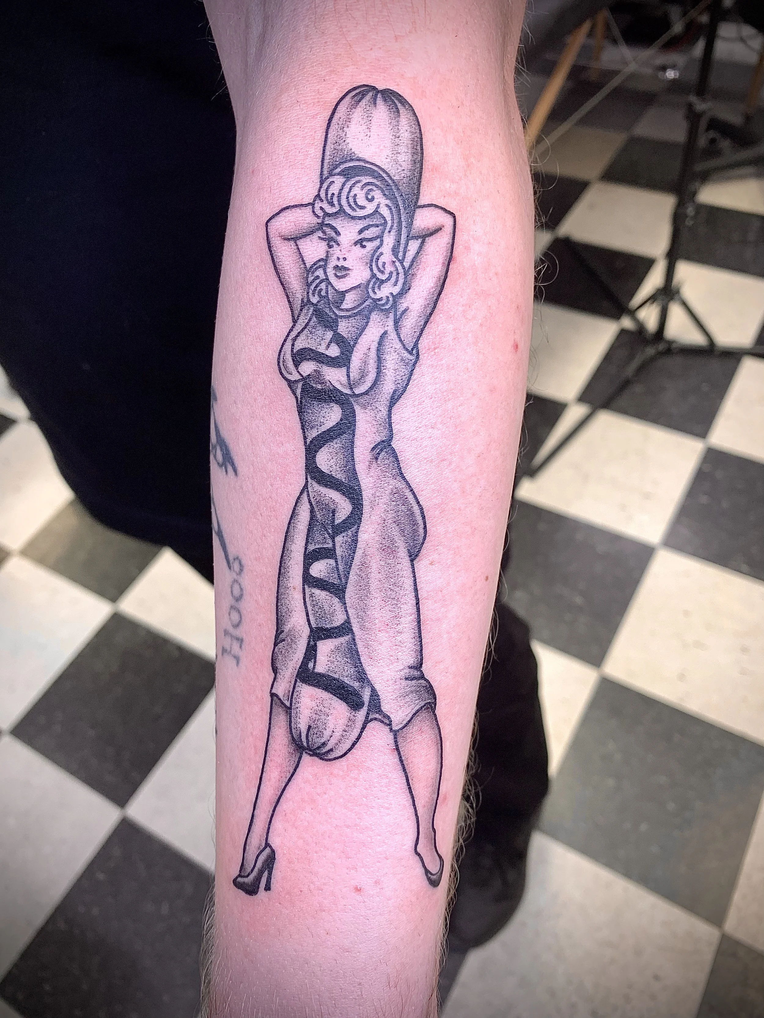 Tattoo of a woman with curly hair, wearing a large hat and high heels, with her arms behind her head.