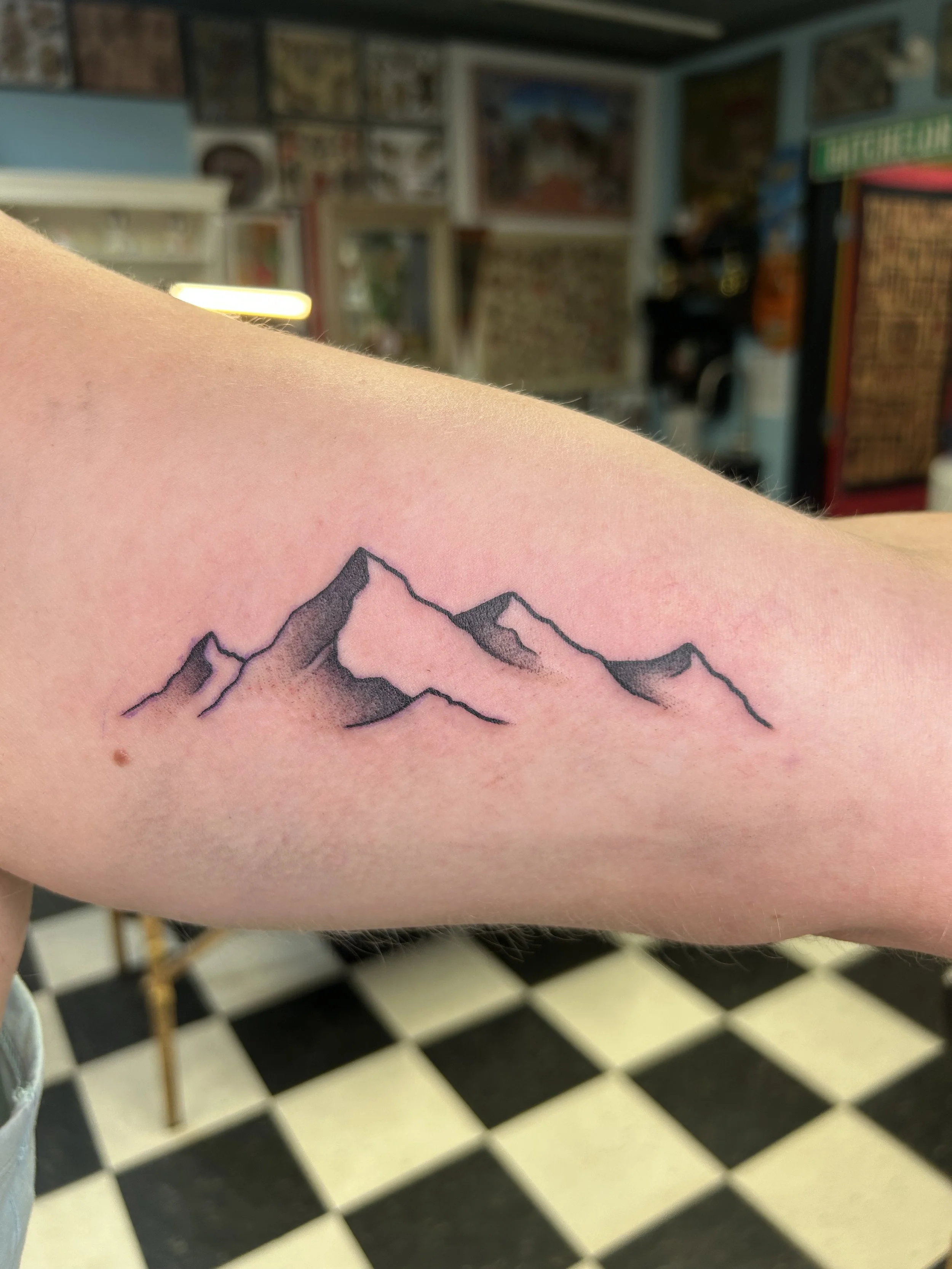 Close-up of a person's arm with a mountain tattoo, with a tattoo shop background and black-and-white checkered floor.
