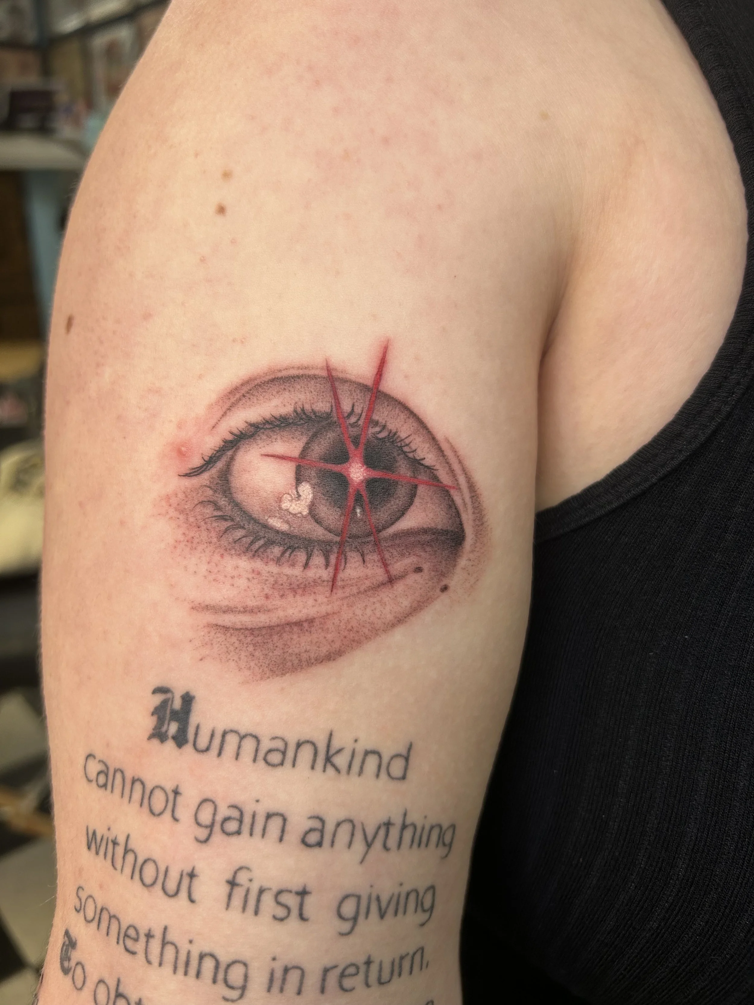 Close-up of a tattoo on a person's arm featuring an eye with detailed eyelashes, and red lines crossing over the eye. Below the tattoo, there is a partially visible text that reads: 'humankind cannot gain anything without first giving.'