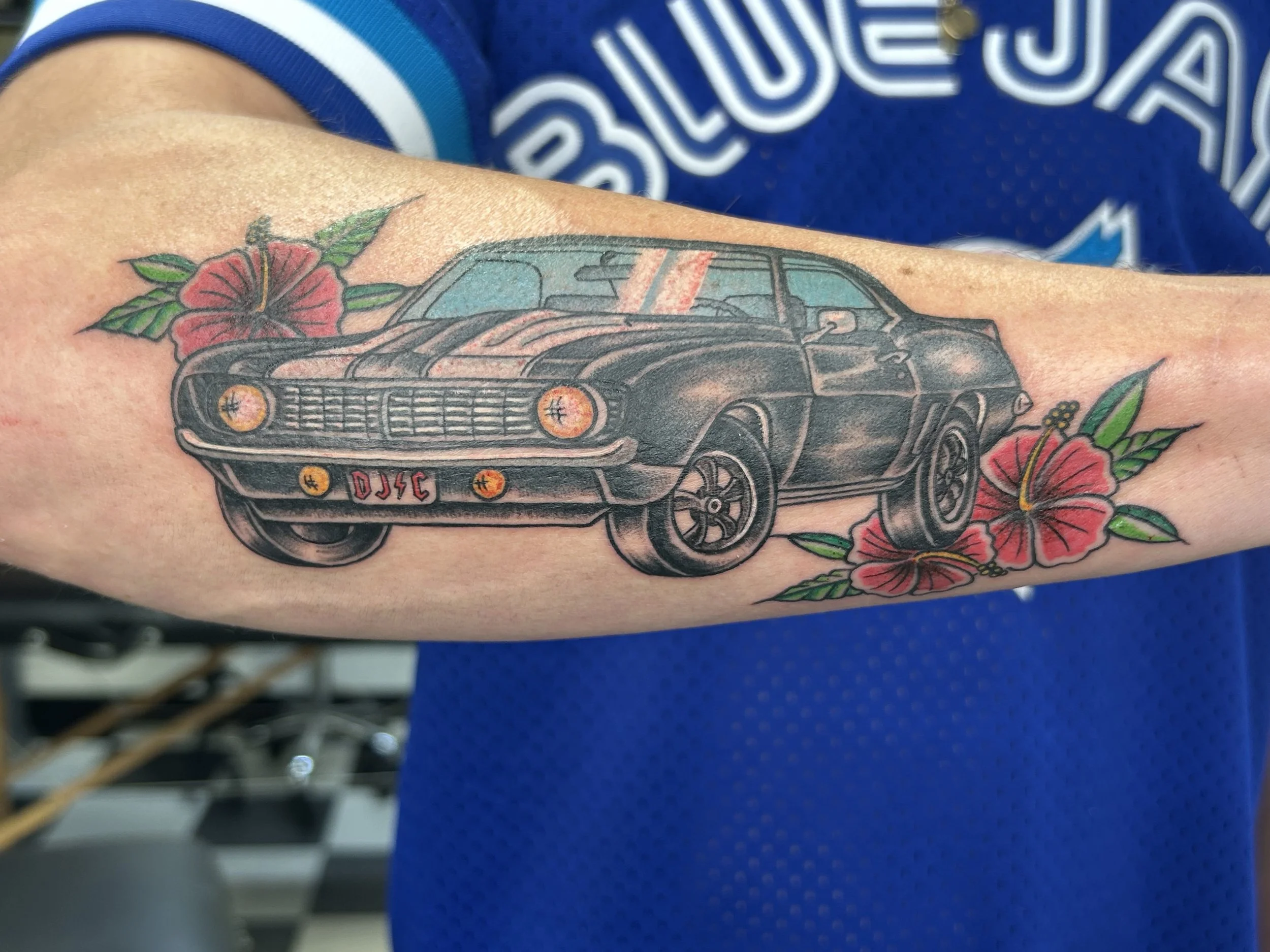 A tattoo of a black muscle car with orange headlights, surrounded by red and pink hibiscus flowers and green leaves on someone's arm, with a blue shirt in the background.
