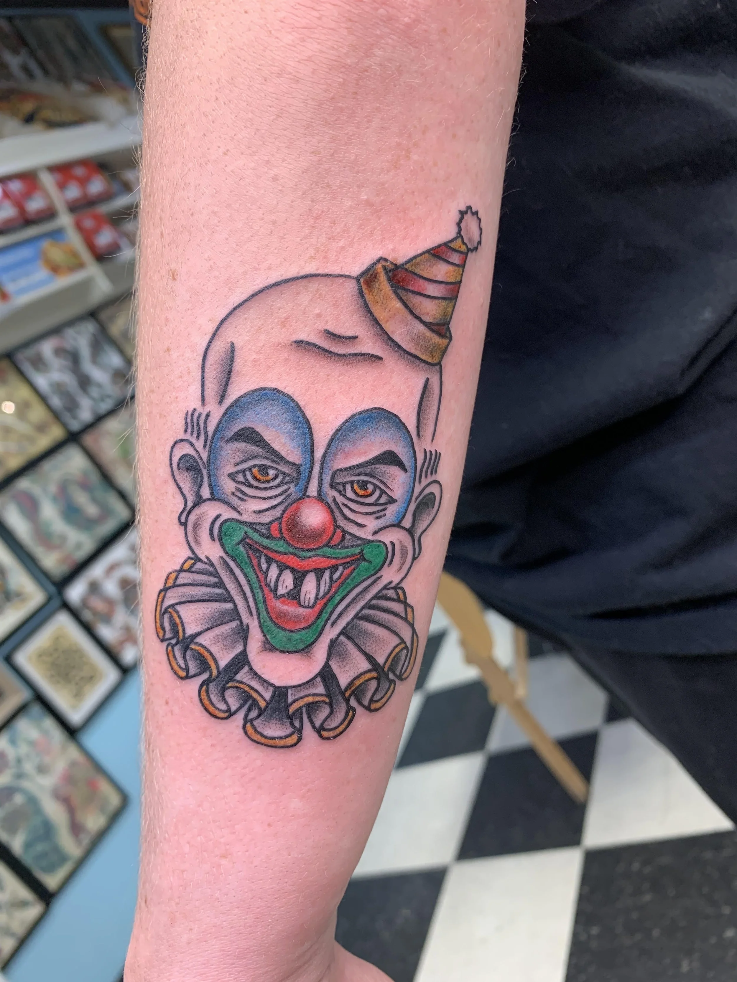 Tattoo of a clown with a colorful hat, exaggerated facial features, and a big red nose on a person's arm.