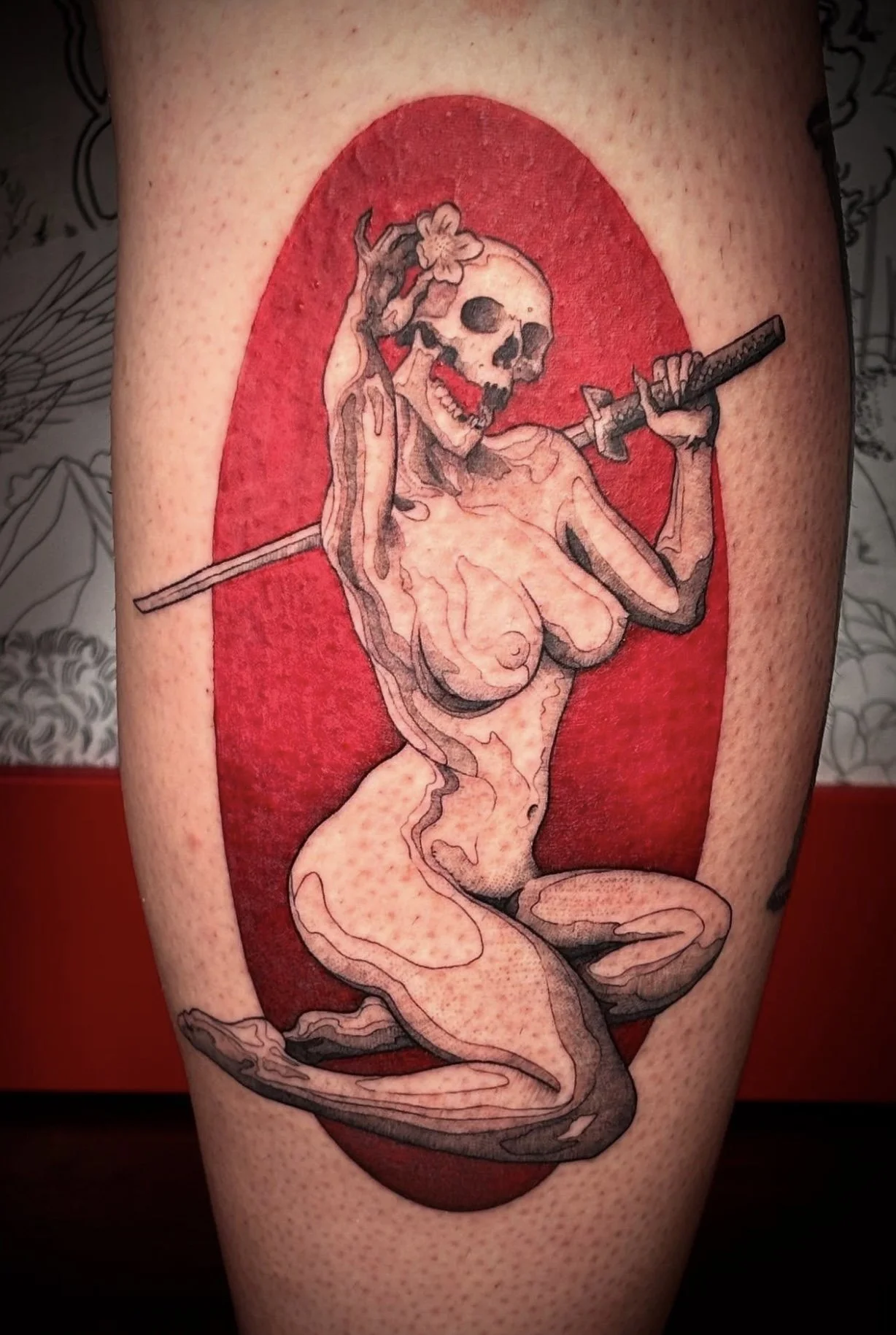 Tattoo of a female skeleton holding a sword with a flower in her hair, set against a red oval background.