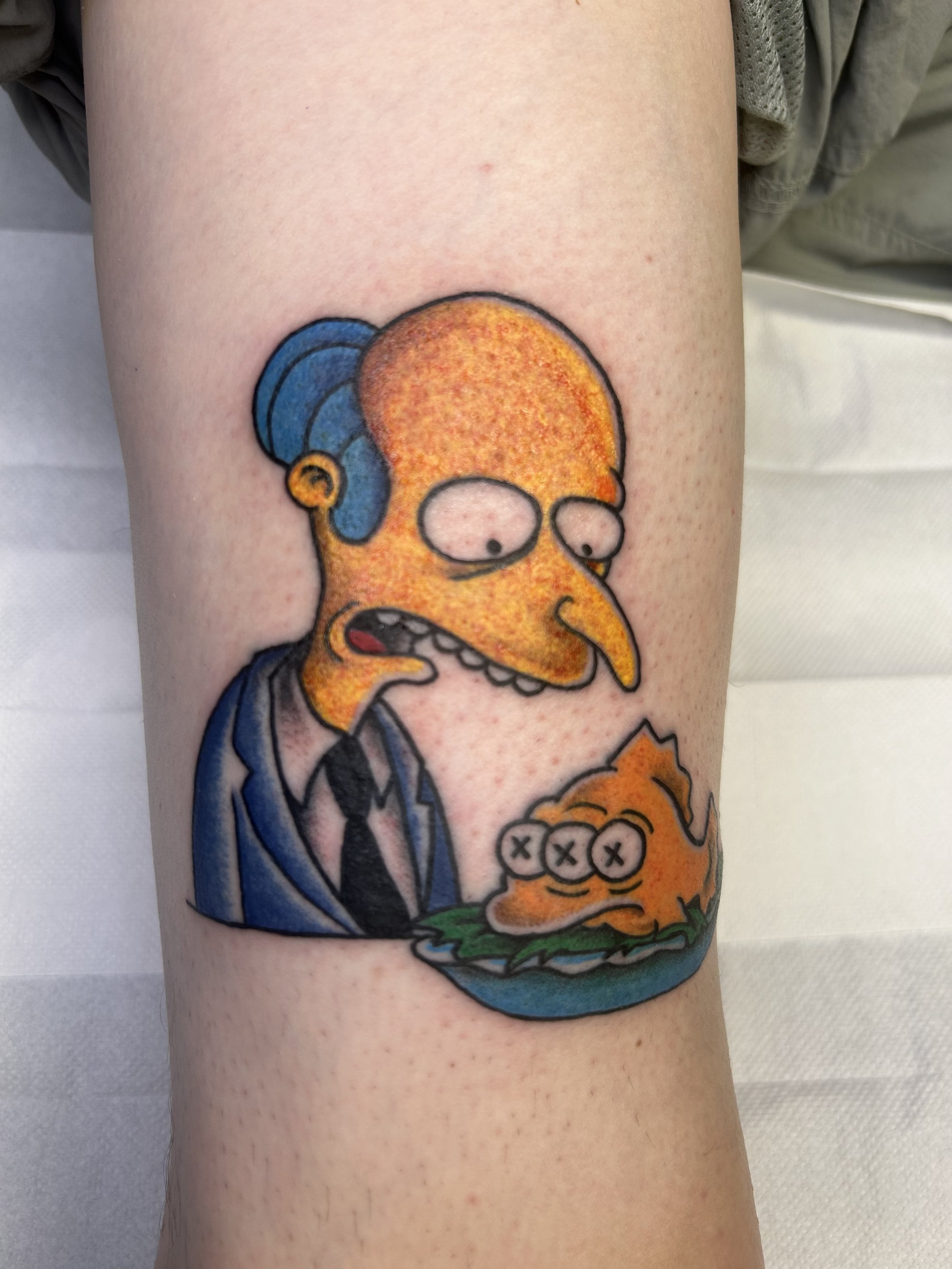 Colorful tattoo of a cartoonish character resembling a bird with an orange beak, blue hair, and a striped blue and gray shirt on a person's arm.