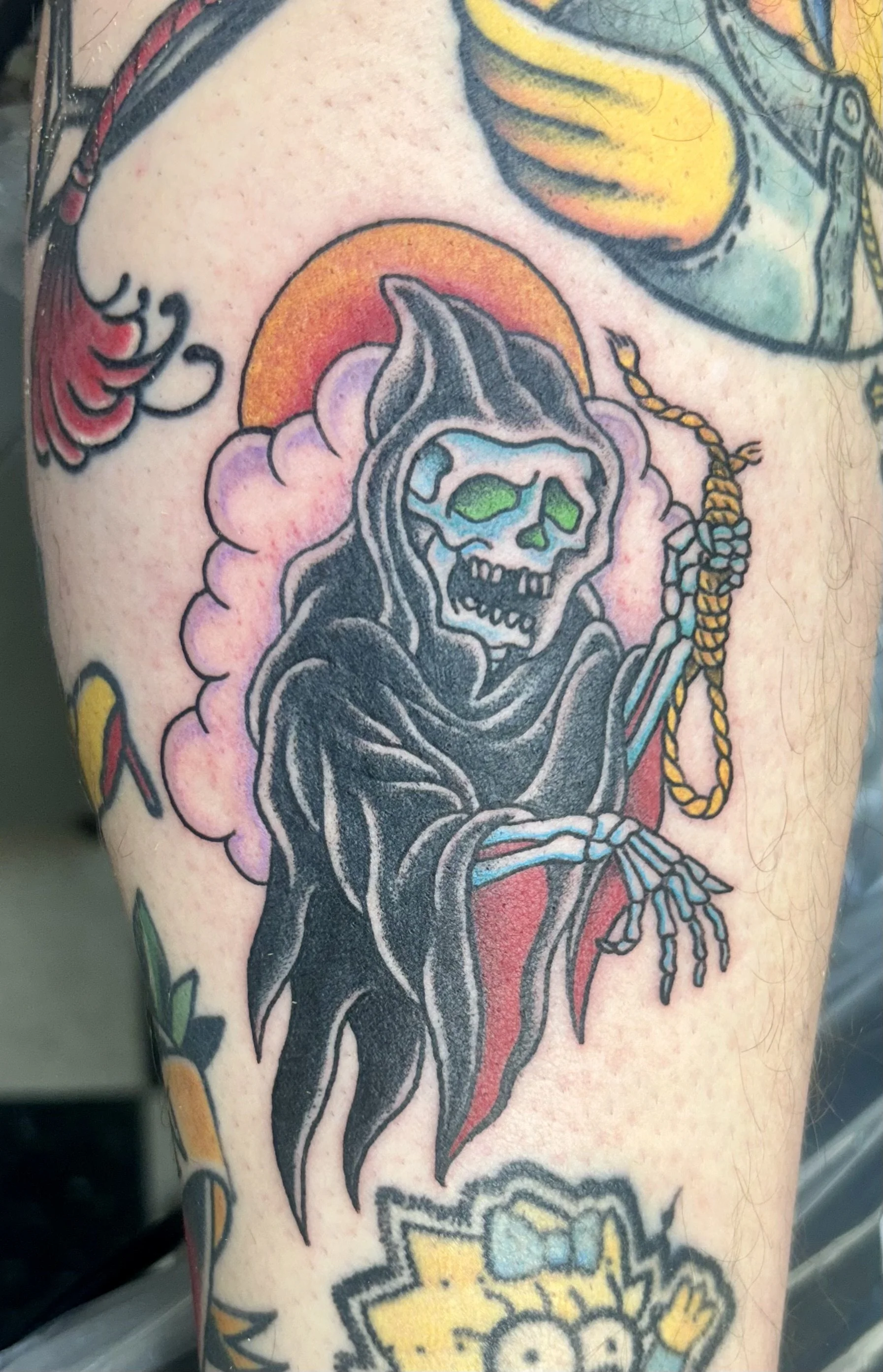 Tattoo of a grim reaper with green eyes and skull face, holding a rope with a sunset and clouds in the background.