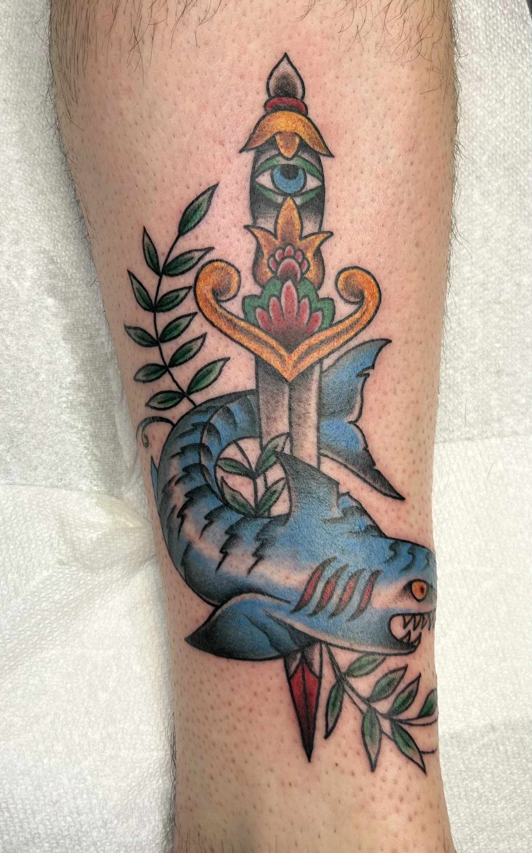 Tattoo of a blue fish with green leaves on the side, a sword with a decorative hilt in the center, and a candle with a flame on top. The fish has red and orange accents and is detailed with shading and outlines.