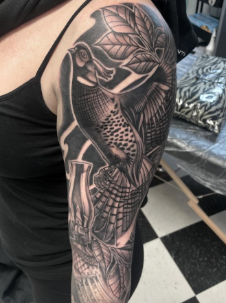 Black and gray tattoo of a toucan perched on a branch with large tropical leaves on the upper arm.