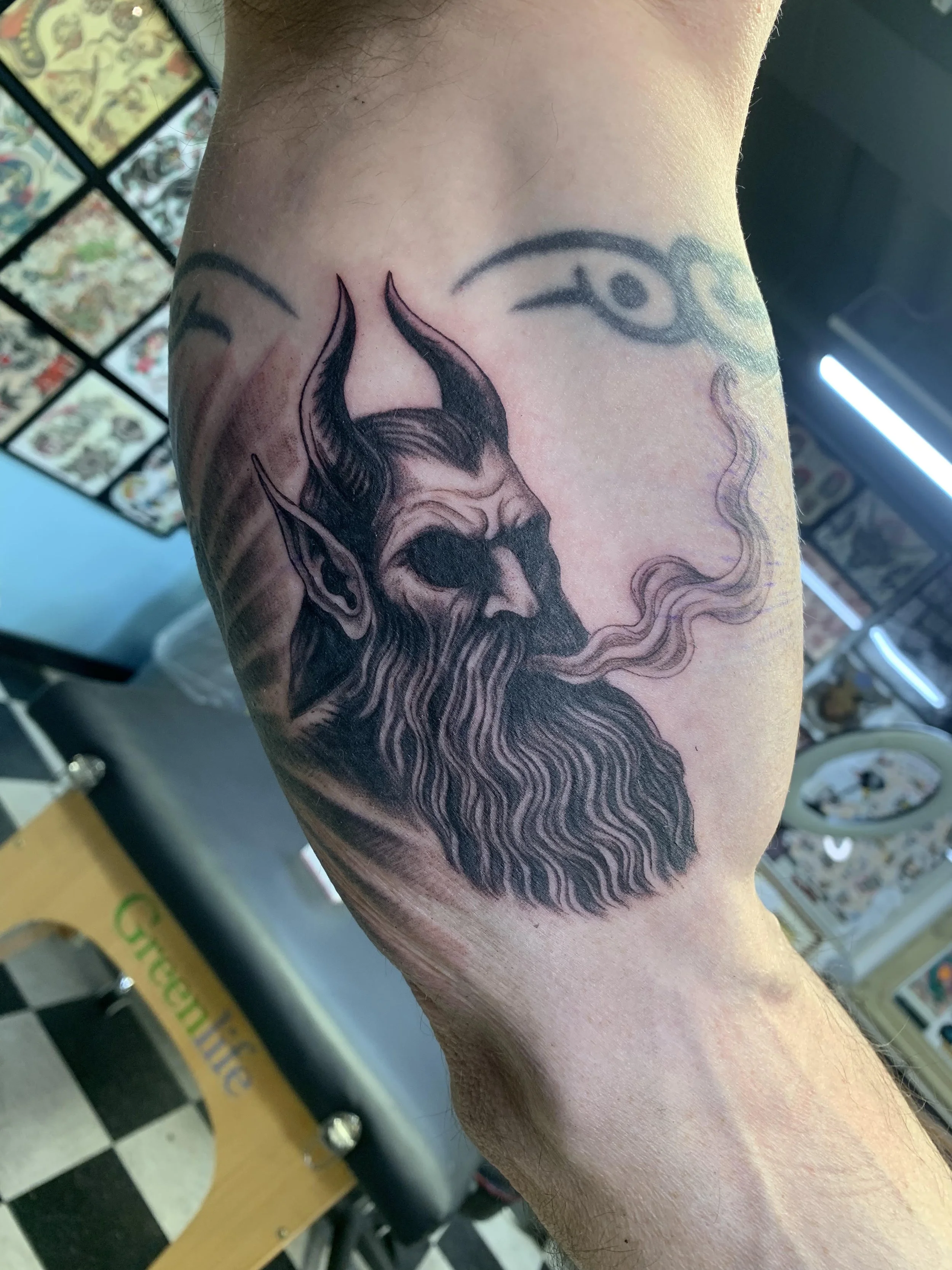 Tattoo of a bearded figure with horns or flames on their head, smoking, and stylized eye symbols above.