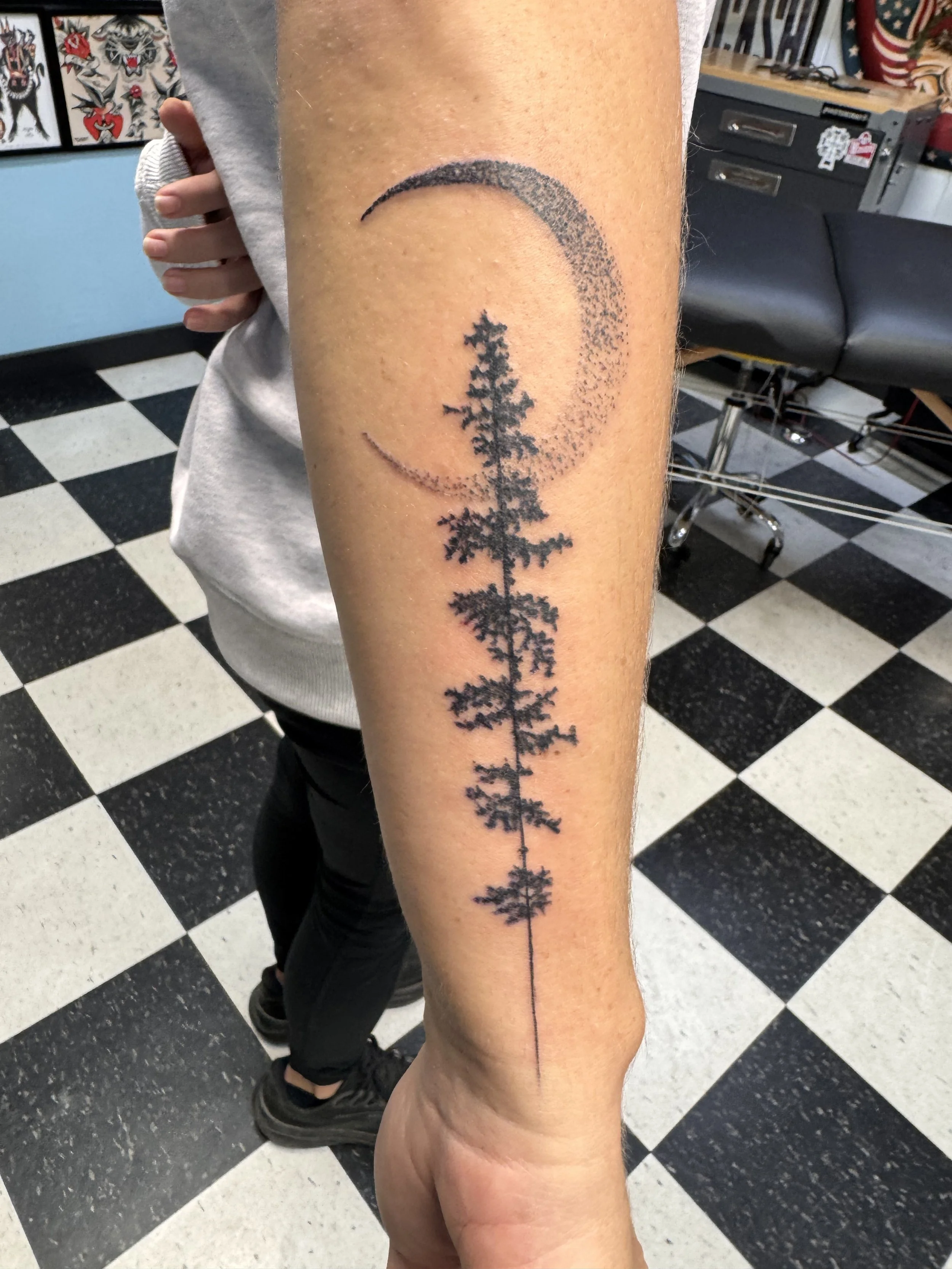 Tattoo of a pine tree within a crescent moon on a person's inner forearm.