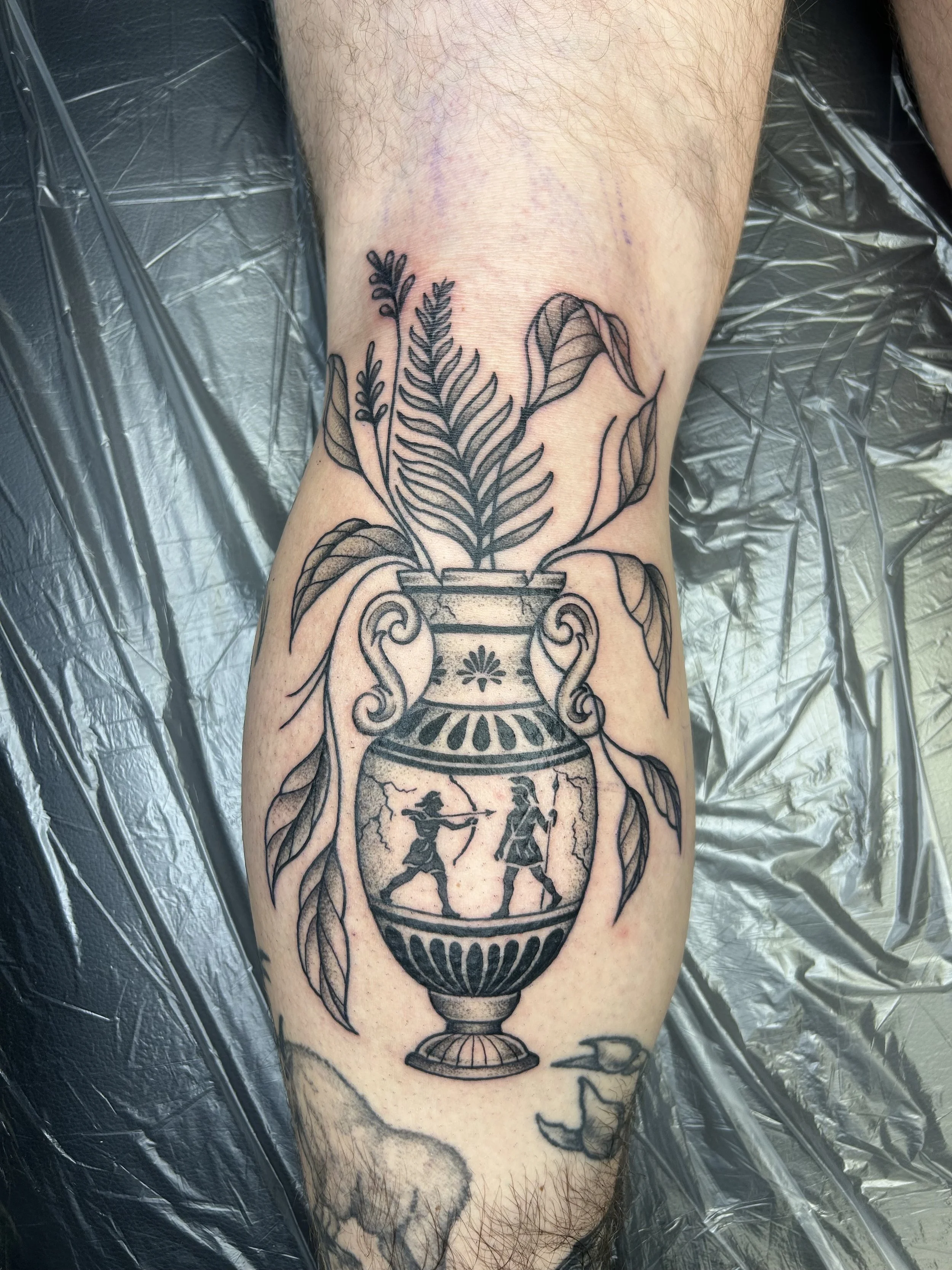 A tattoo of an ornate vase with leaves and plants, with two dancers inside the vase, on a person’s lower leg.