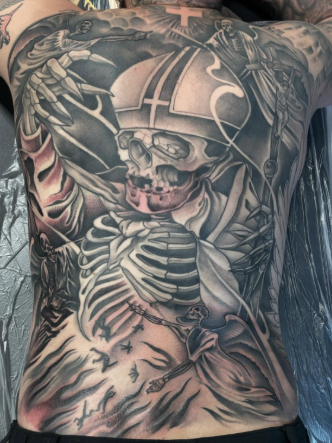 A detailed black and gray tattoo featuring a skull wearing a military helmet with a cross, a skeleton with ribs exposed, and a scorpion, covering the person's chest and abdomen.