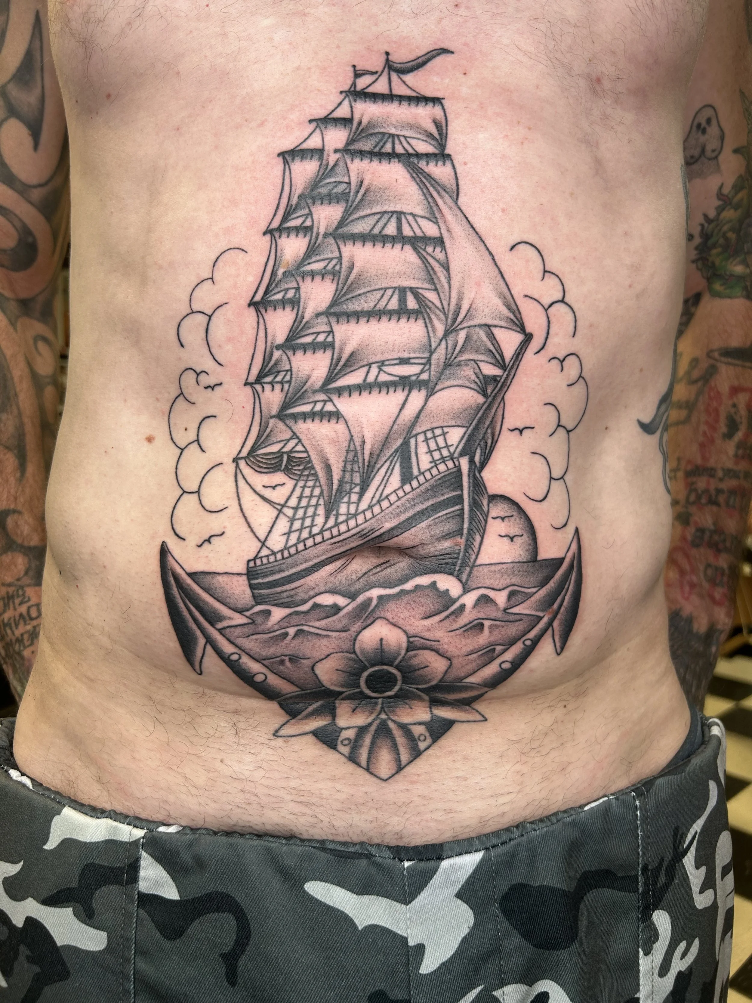 A tattoo of a sailing ship with multiple sails on a person's chest, surrounded by clouds and waves, with tattoos on both arms.