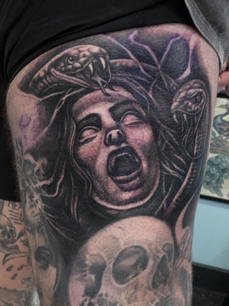 Tattoo of a woman's face with her mouth open, surrounded by a snake with its mouth open and fangs showing, and a skull below.