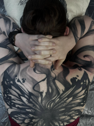 Person with large black and gray tattoos across their back and arms, covering the shoulder blades and extending to the arms, with hands clasped behind their head and short dark hair.